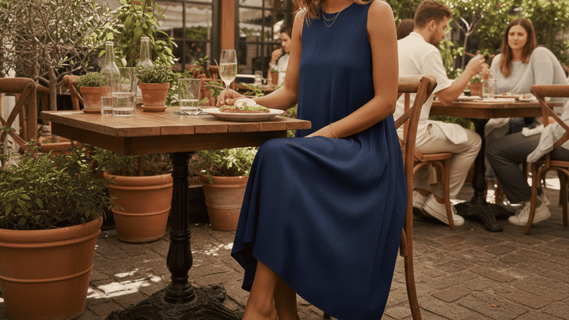Woman in a navy dress and espadrilles enjoys an outdoor brunch under a flowering pergola with a white farmhouse in the background.