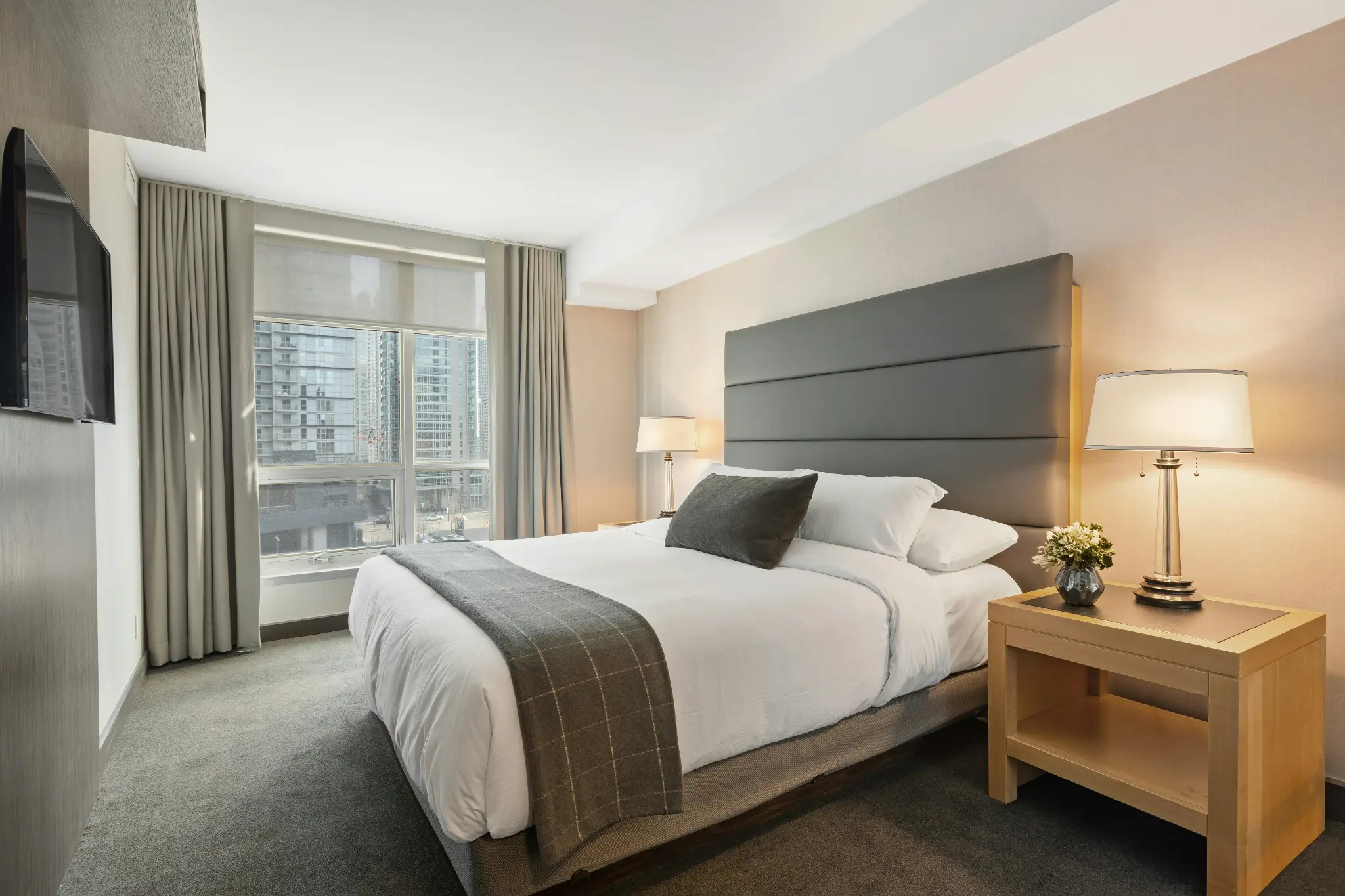 Modern hotel room with a king-size bed, gray headboard, and bedside lamp near a large window