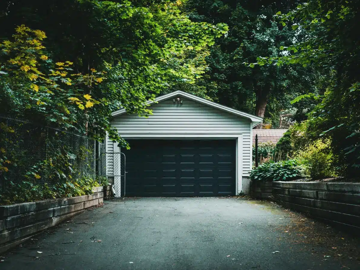 White garage with black door surrounded by lush green foliage in a wooded setting