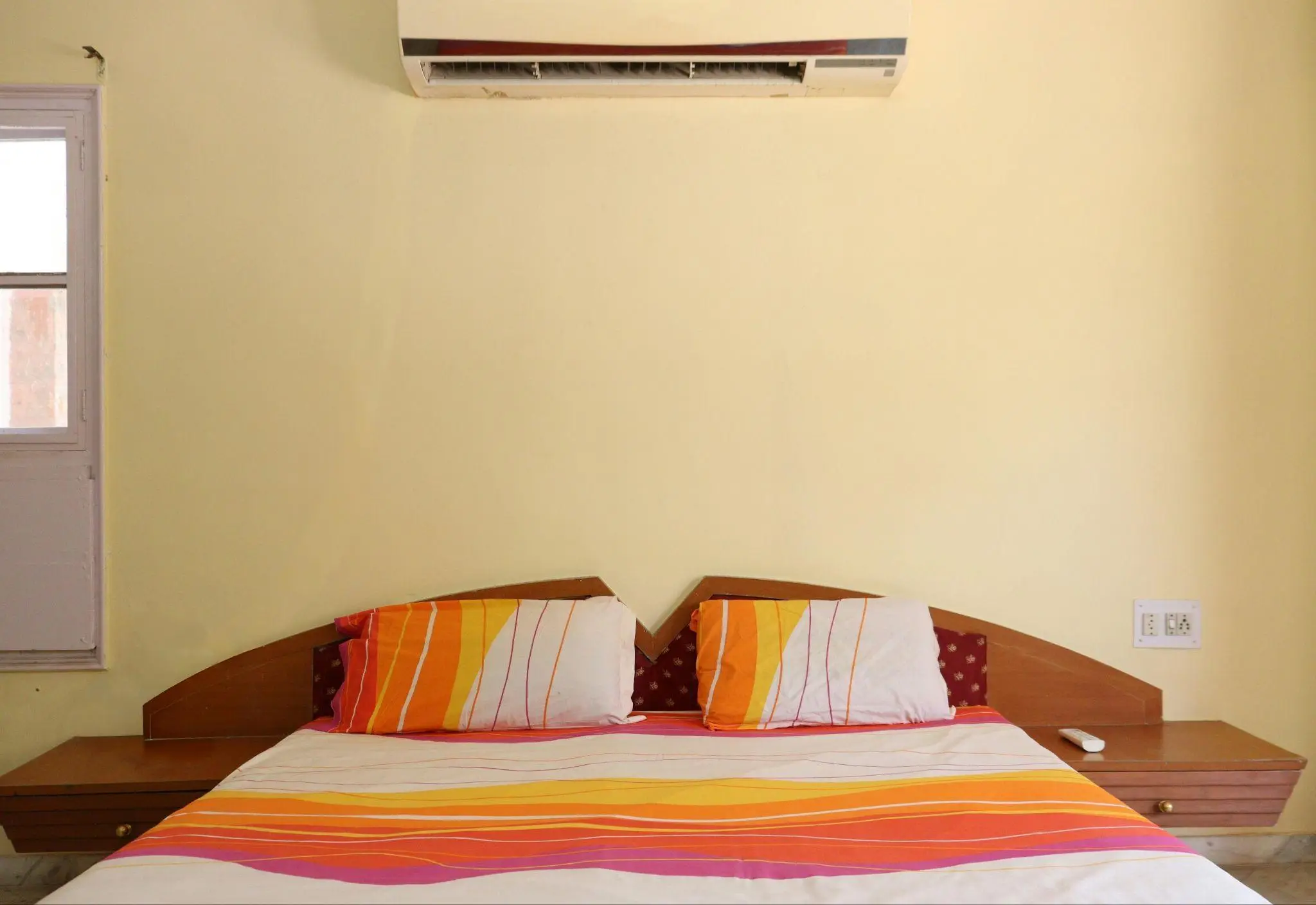 Bed with striped orange bedding under wall-mounted air conditioner in a sunlit bedroom