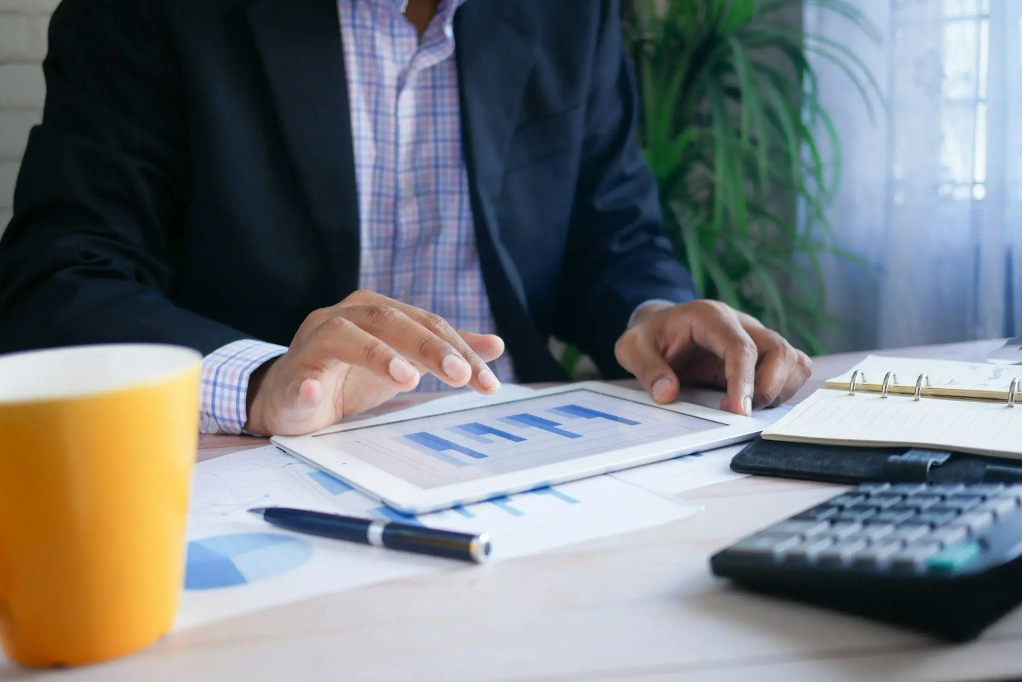 Person in business attire using a tablet with charts at a desk with coffee mug and notebook