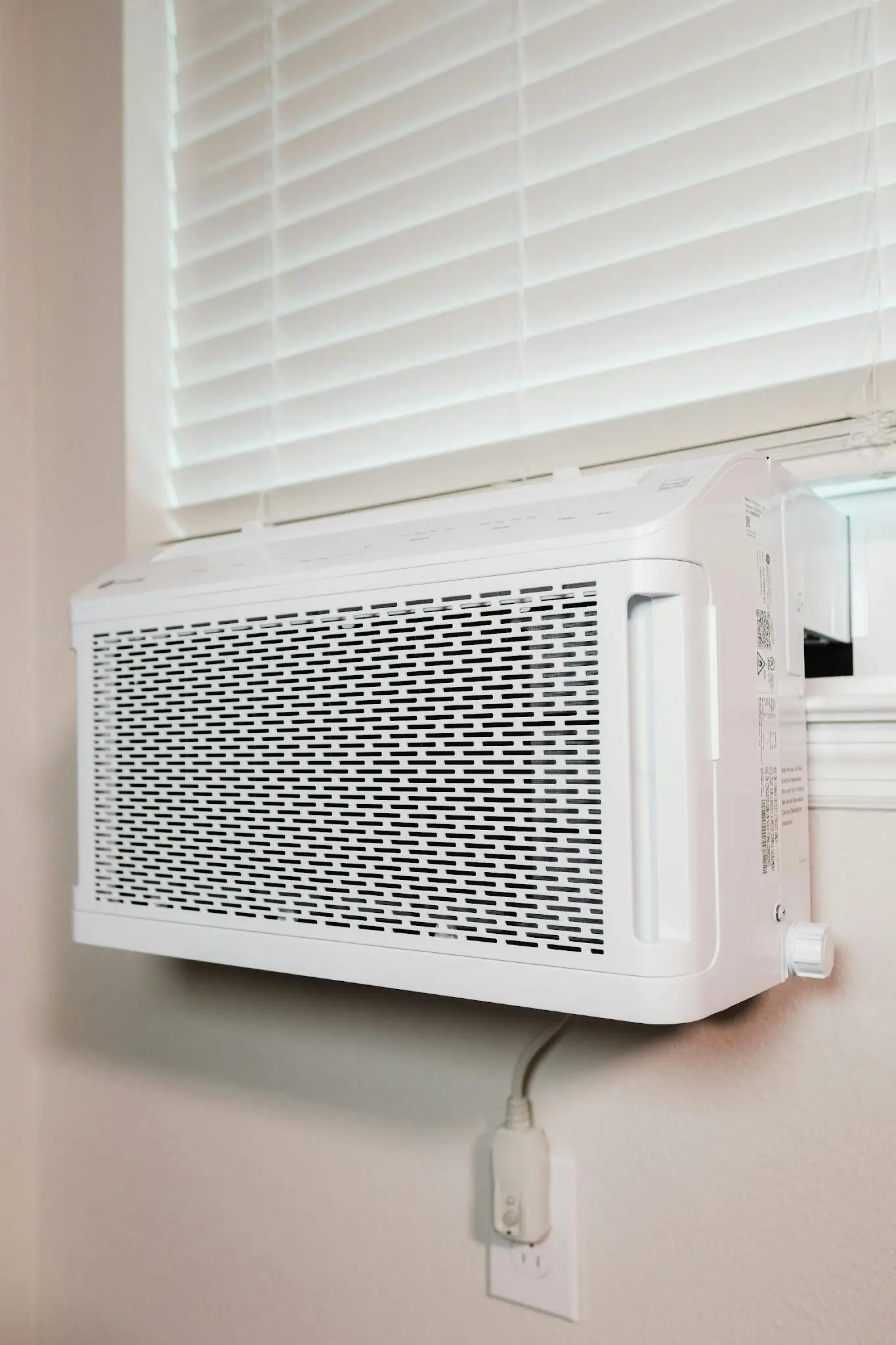 White wall-mounted air conditioner beneath closed blinds in a sunlit indoor setting