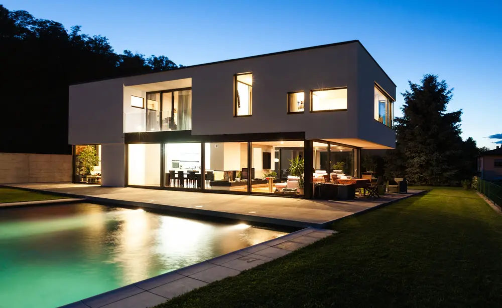 Modern cubic house with illuminated windows beside a pool at dusk in landscaped garden