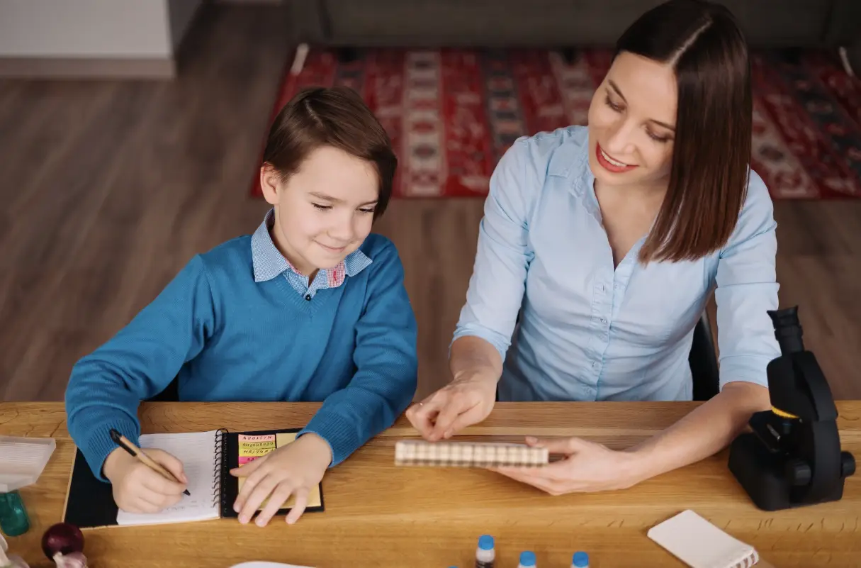 Child writing in notebook with adult in living room, microscope on wooden table