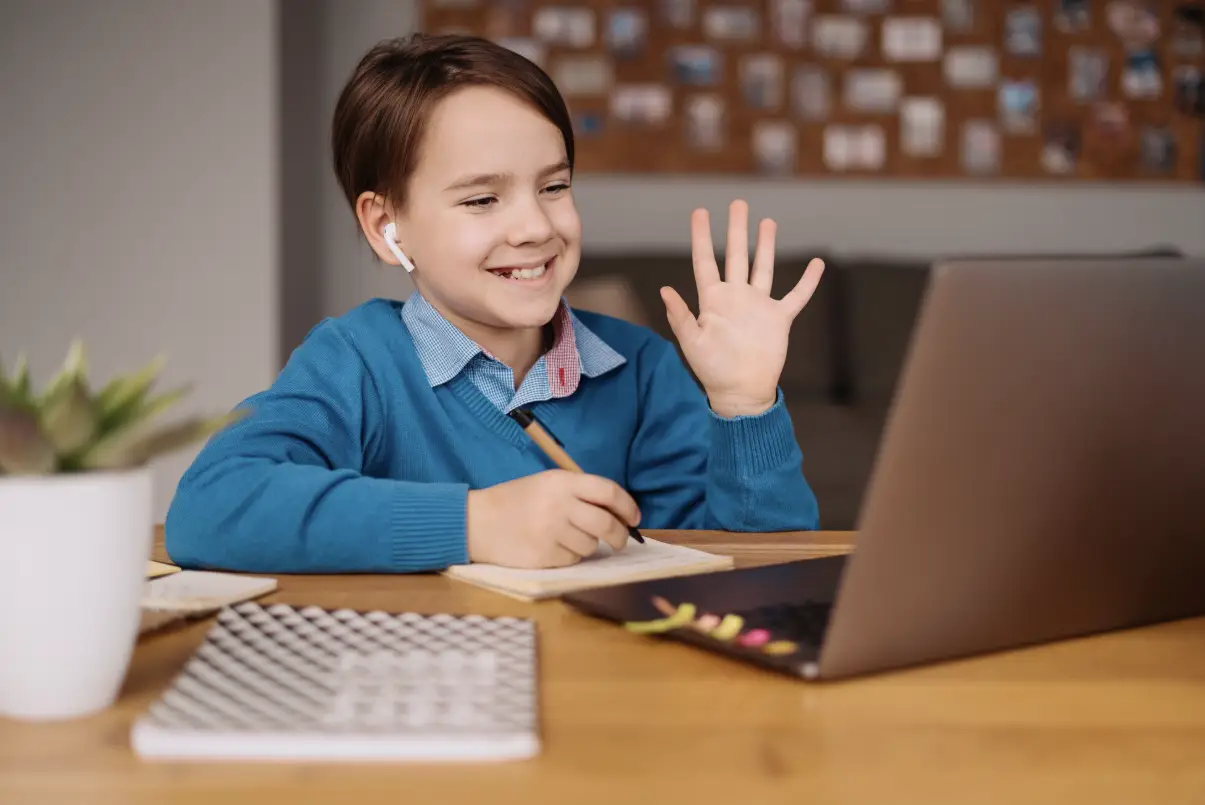Child in blue sweater waving during online class on laptop at home desk