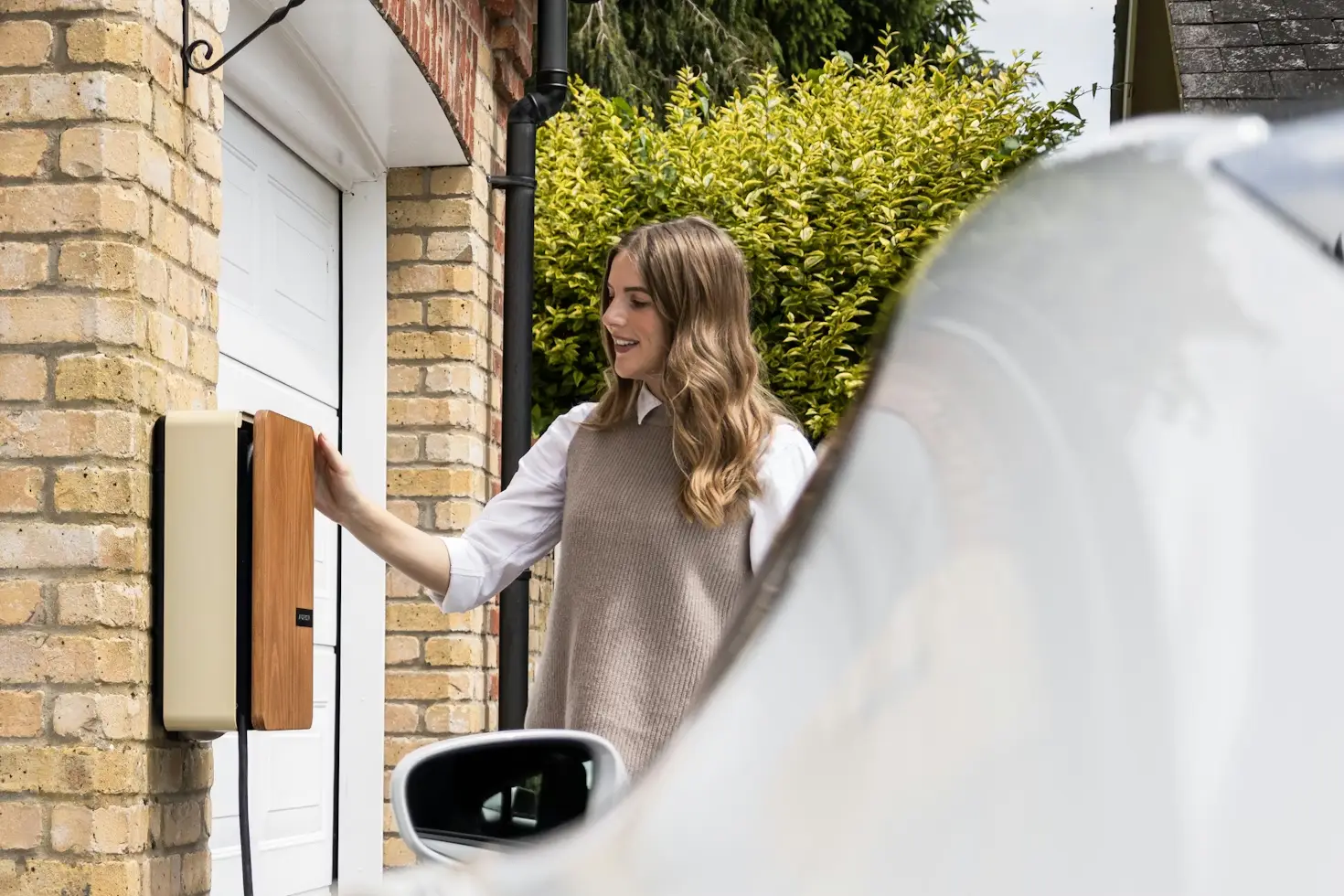 Woman charging electric car at home charging station in driveway with brick wall background