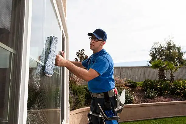 Window cleaner in blue uniform cleaning large glass pane in sunny garden setting