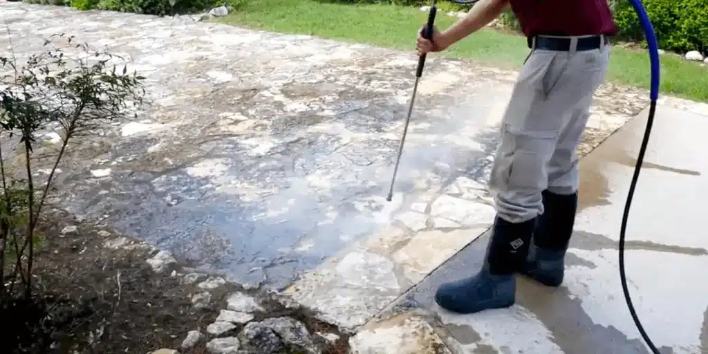 Person using a pressure washer on stone patio outdoors with greenery in background