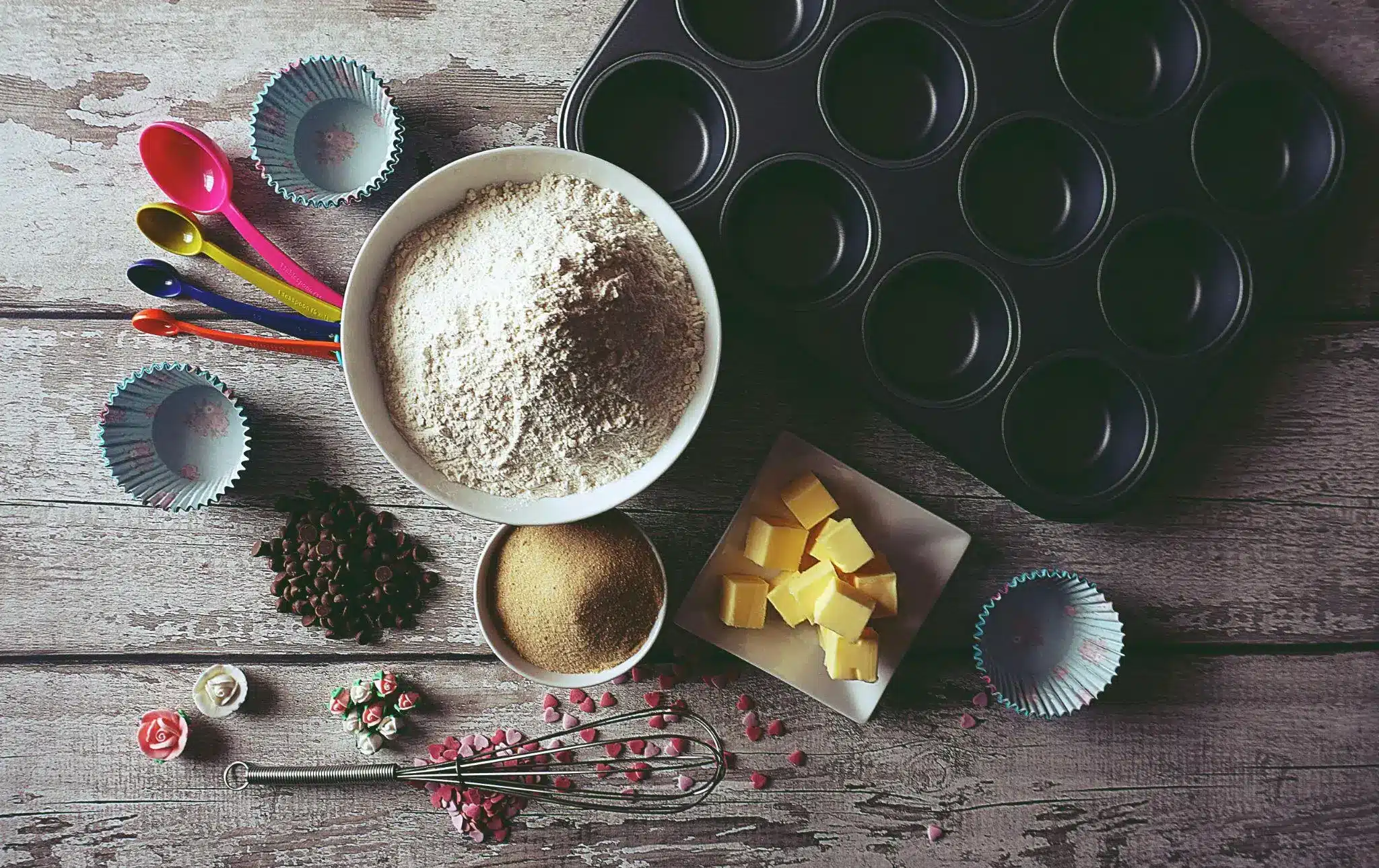 Baking ingredients with flour, butter, and chocolate chips on wooden surface beside muffin tin