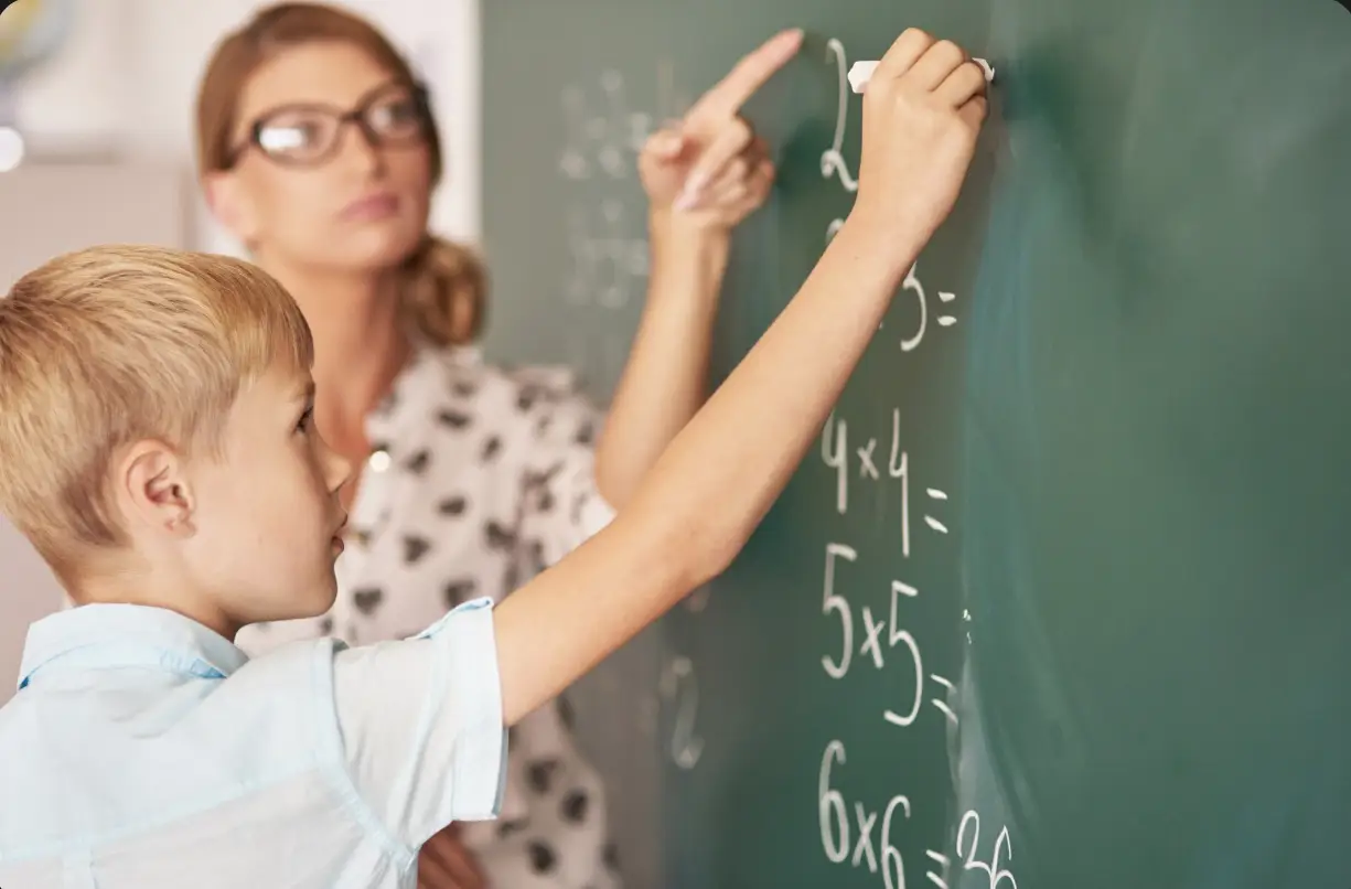 Boy solving math equations on chalkboard with teacher's guidance in classroom setting
