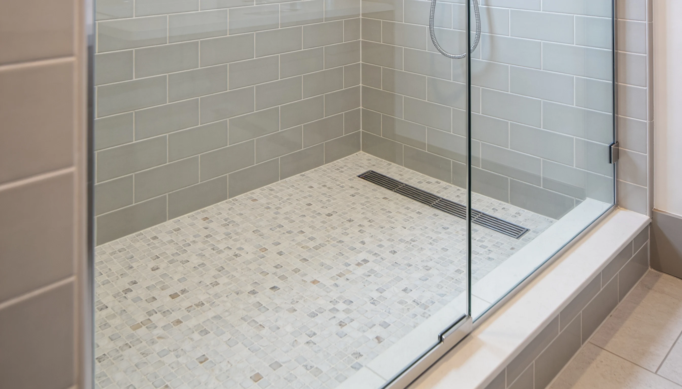 A modern walk-in shower featuring gray subway tile walls a small square mosaic tile floor and a long linear metal drain behind a clear glass enclosure
