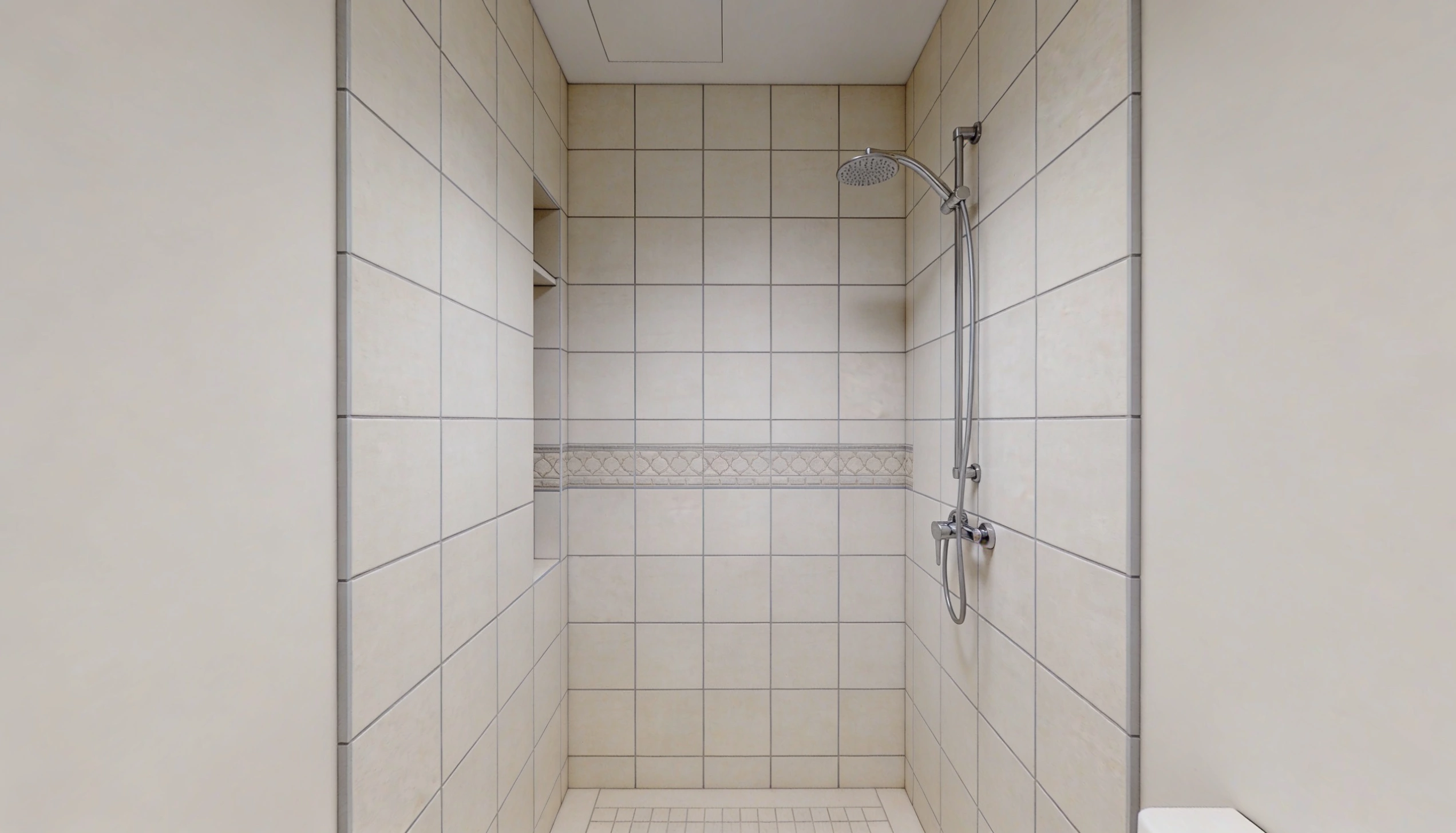 A narrow shower stall featuring beige square tiles a chrome showerhead a recessed shelf and contrasting white outer walls