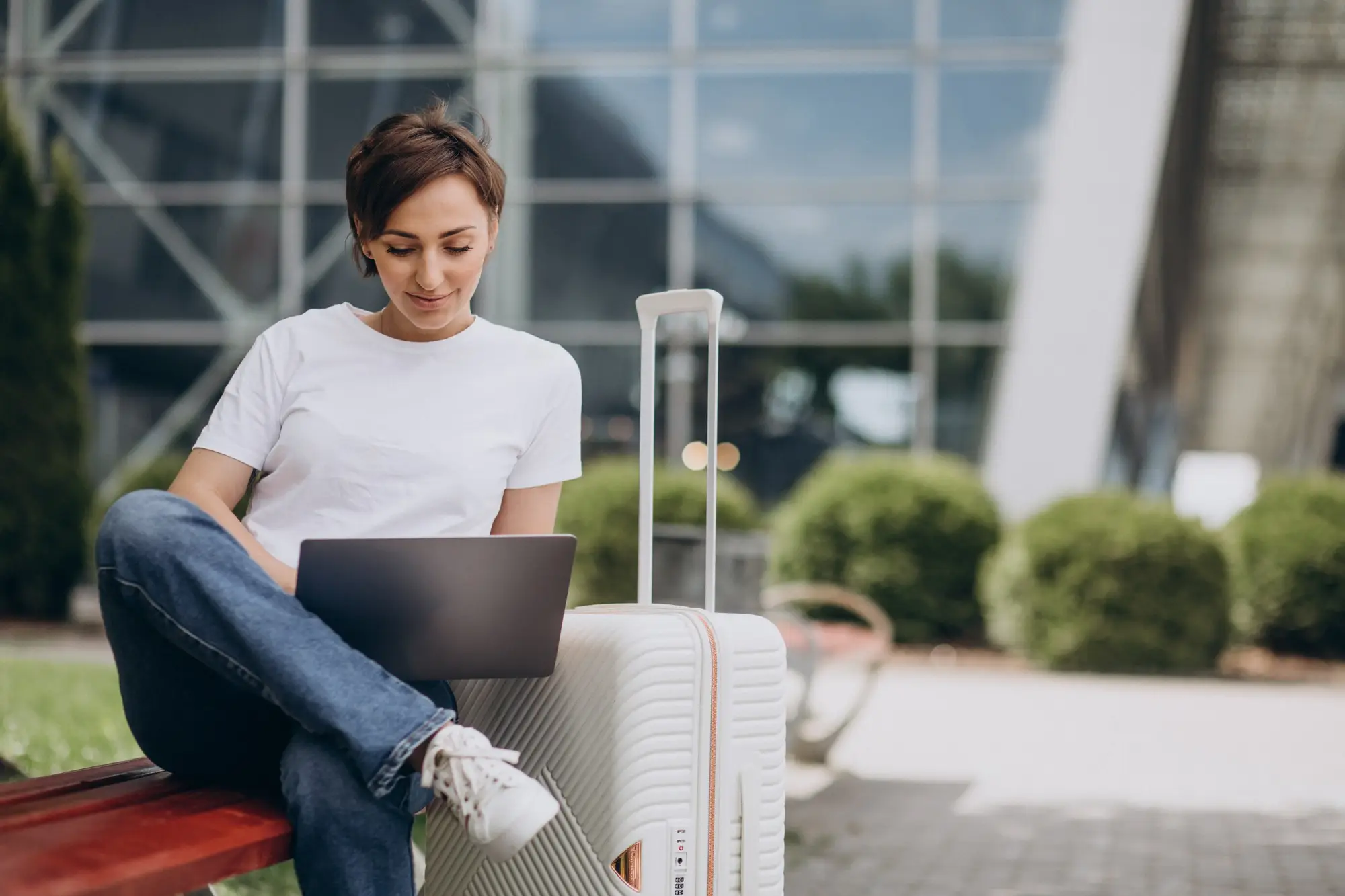 Woman in white t-shirt using laptop next to suitcase outside modern building