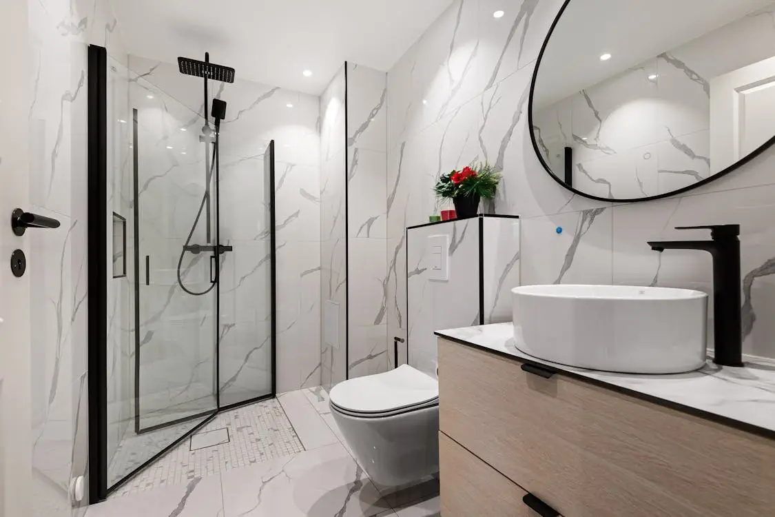 Modern bathroom with glass shower, circular mirror, and marble vanity in a bright setting
