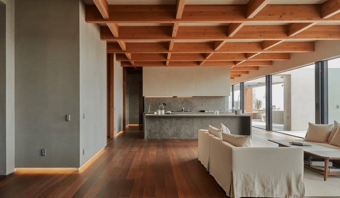 Modern living room with warm wood floors and ceiling beams, smooth concrete kitchen, soft furniture, and large glass windows.