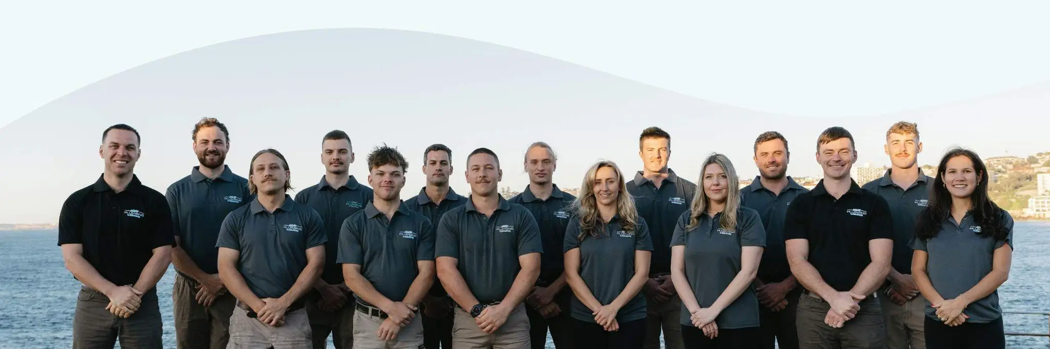 Group of people in matching dark blue shirts standing outdoors with water and buildings in background