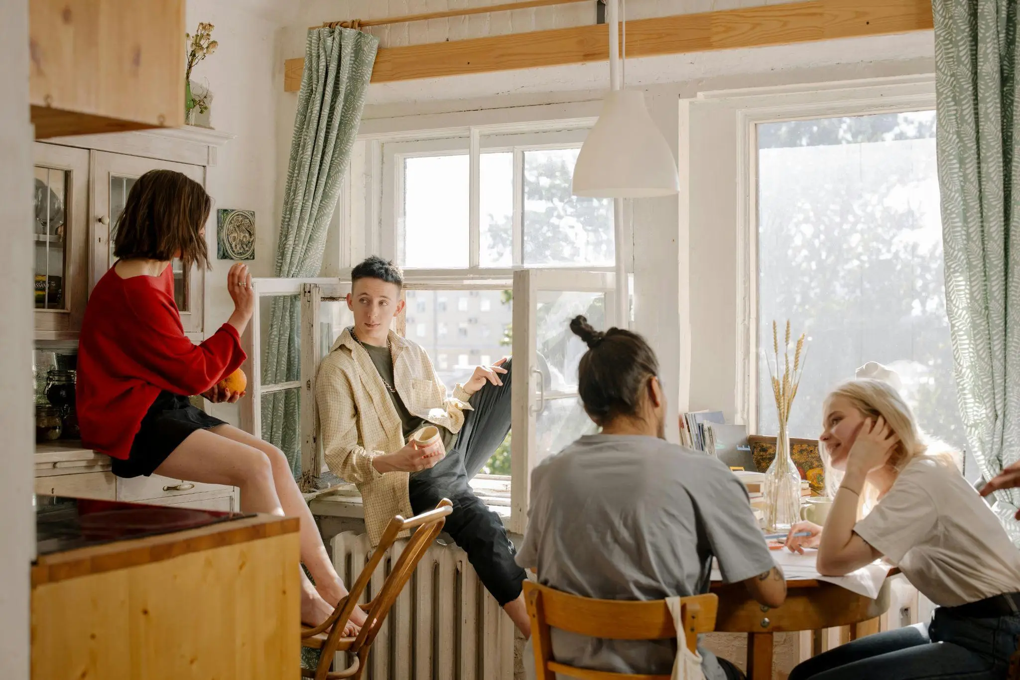 Group of people socializing in a sunlit kitchen with wooden furniture and green curtains