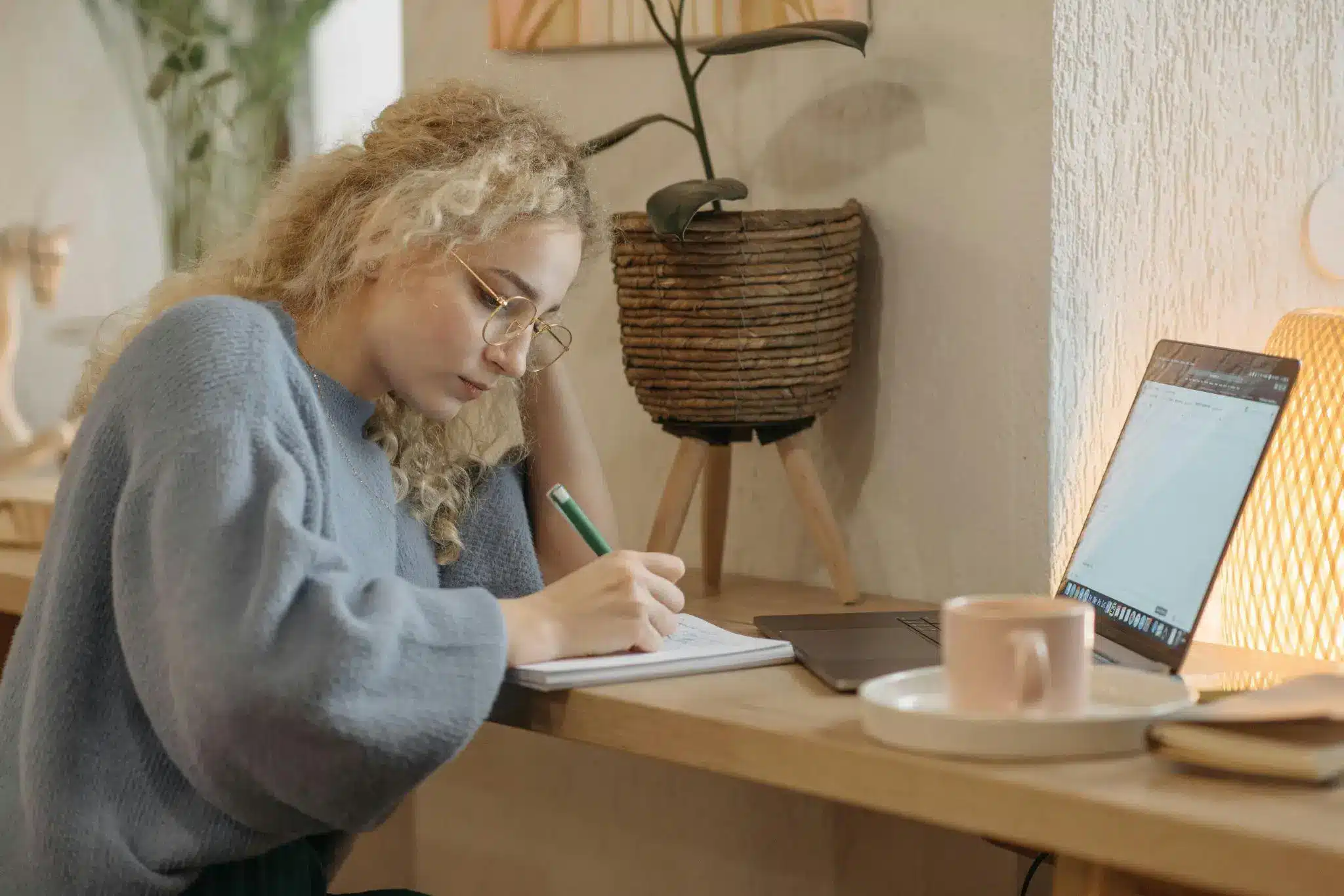Young woman with curly hair writing in notebook at cozy wooden desk with laptop and plant