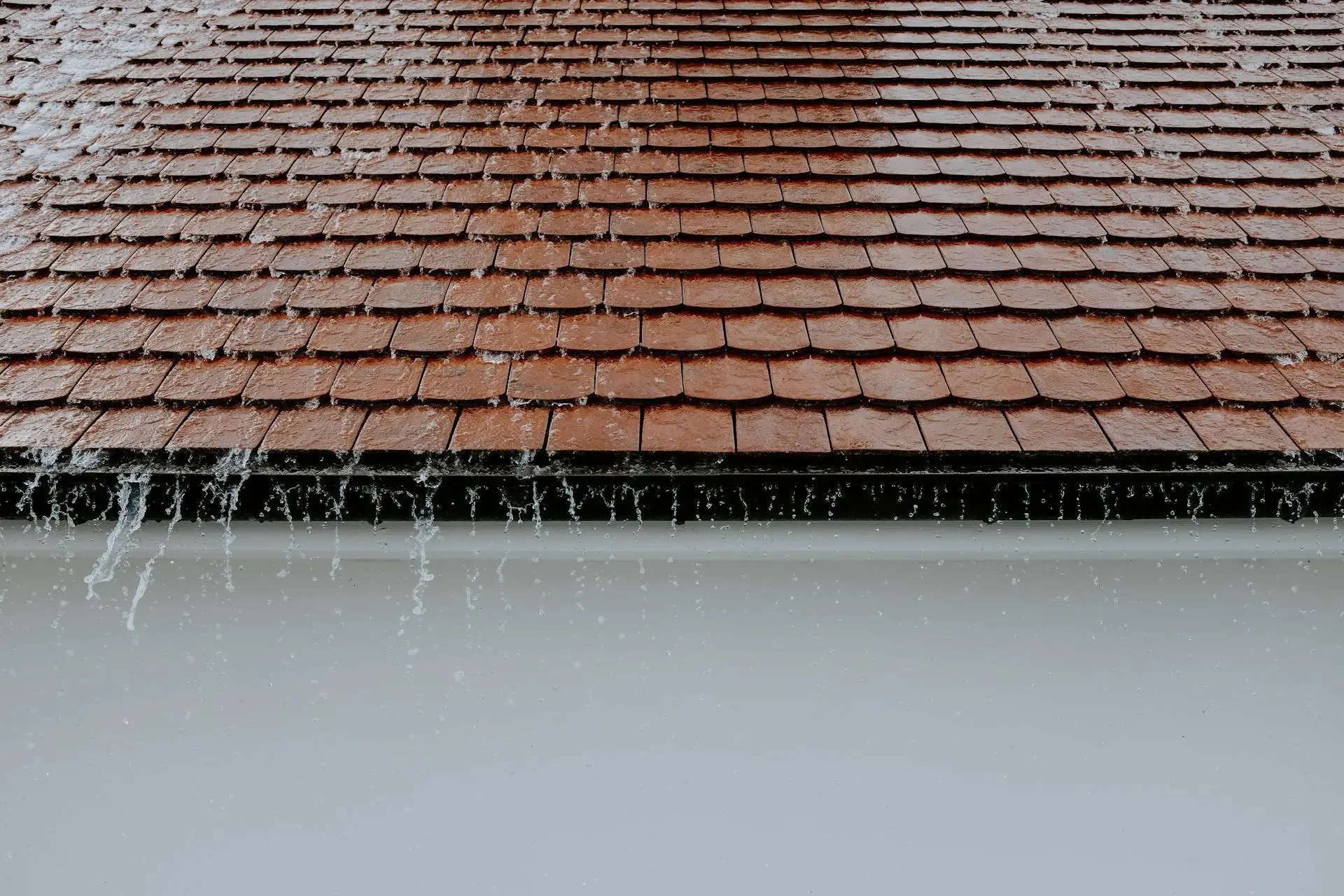 Rain pouring off brown shingle roof onto white wall in outdoor setting