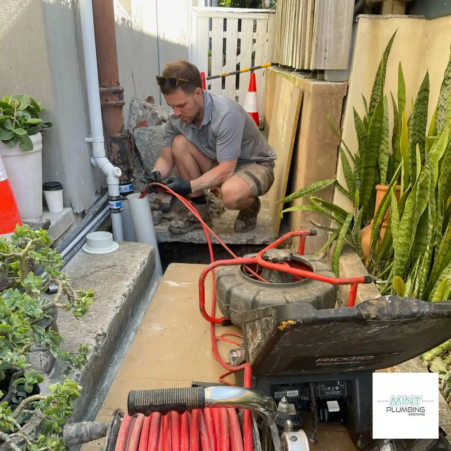Plumber working on outdoor pipe repair with tools and plants nearby