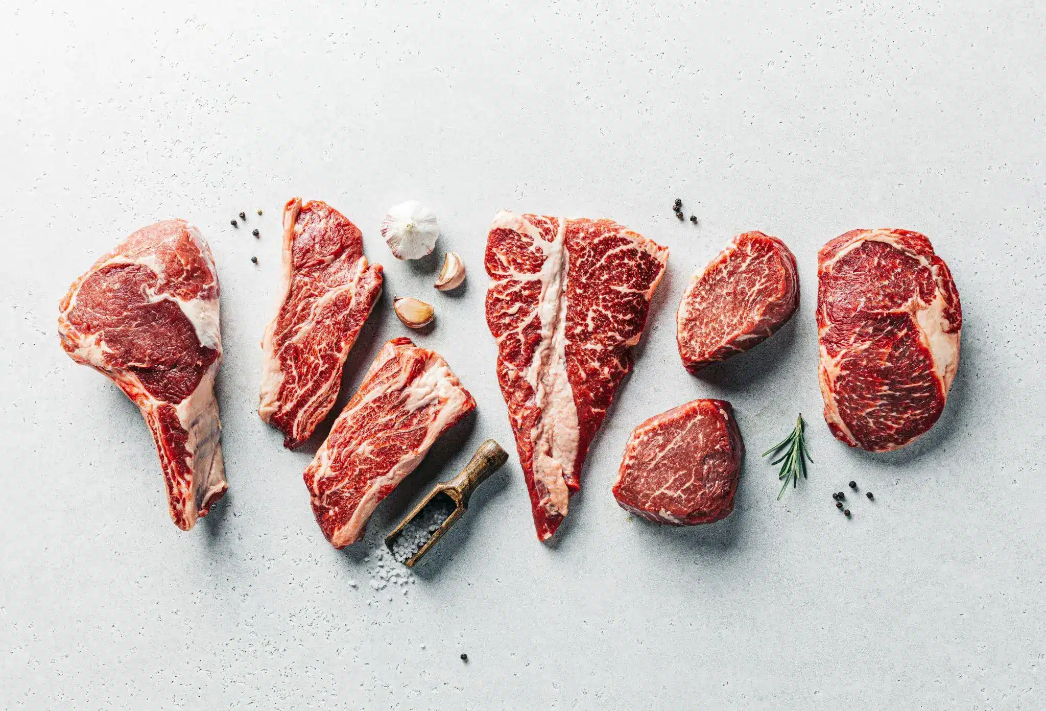 Various cuts of raw steak arranged with garlic and peppercorns on light gray surface