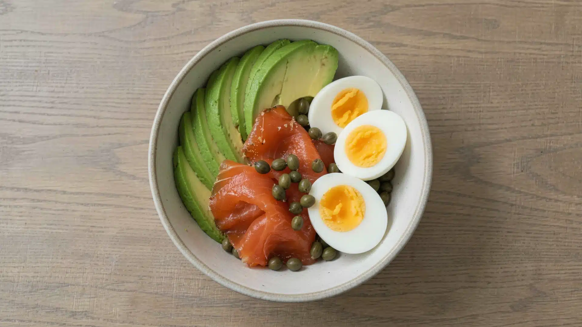 Bowl of sliced avocado, smoked salmon, hard boiled eggs, and capers on a wooden table