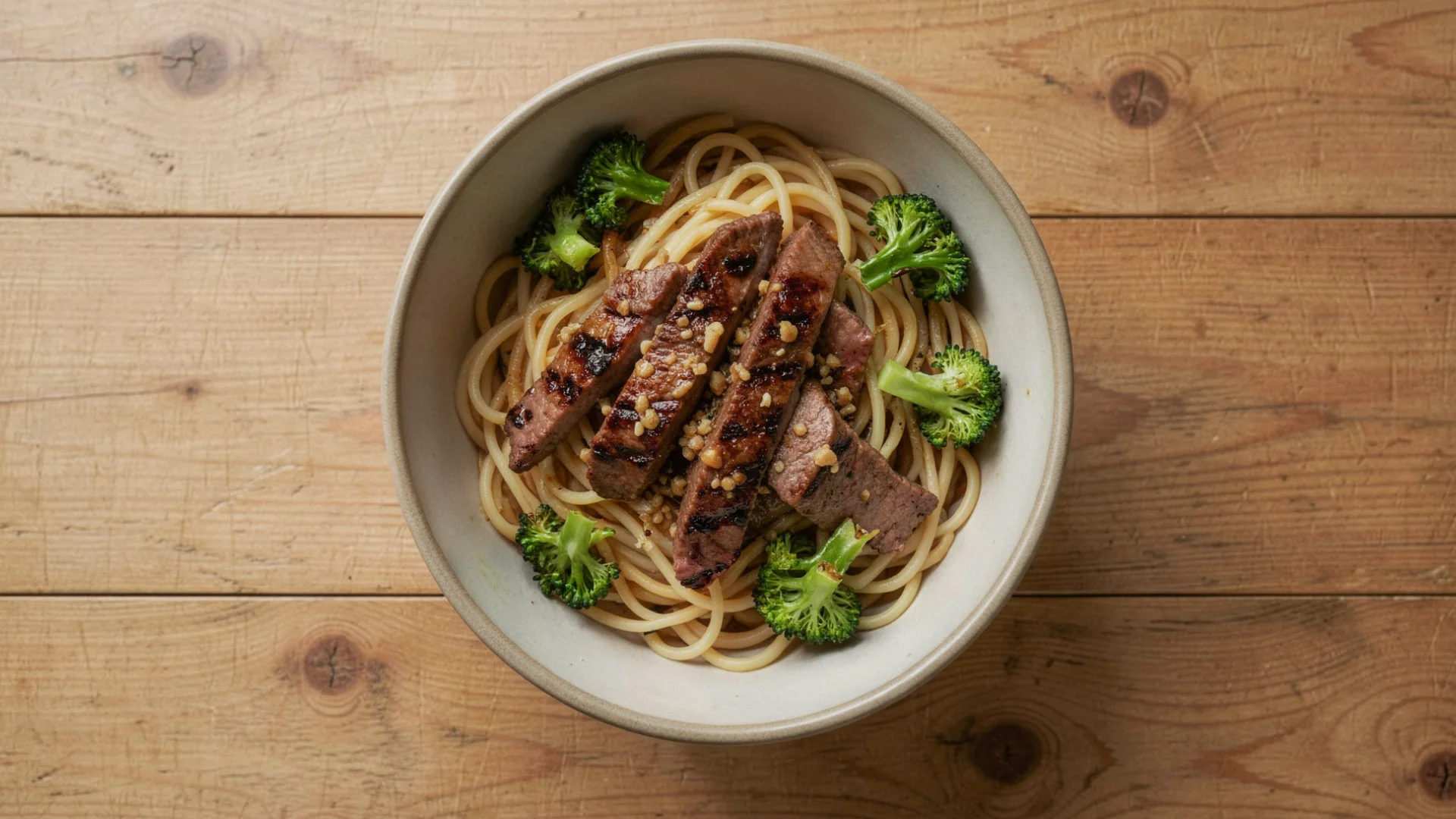 Bowl of spaghetti topped with sliced grilled steak and broccoli florets on a wooden table