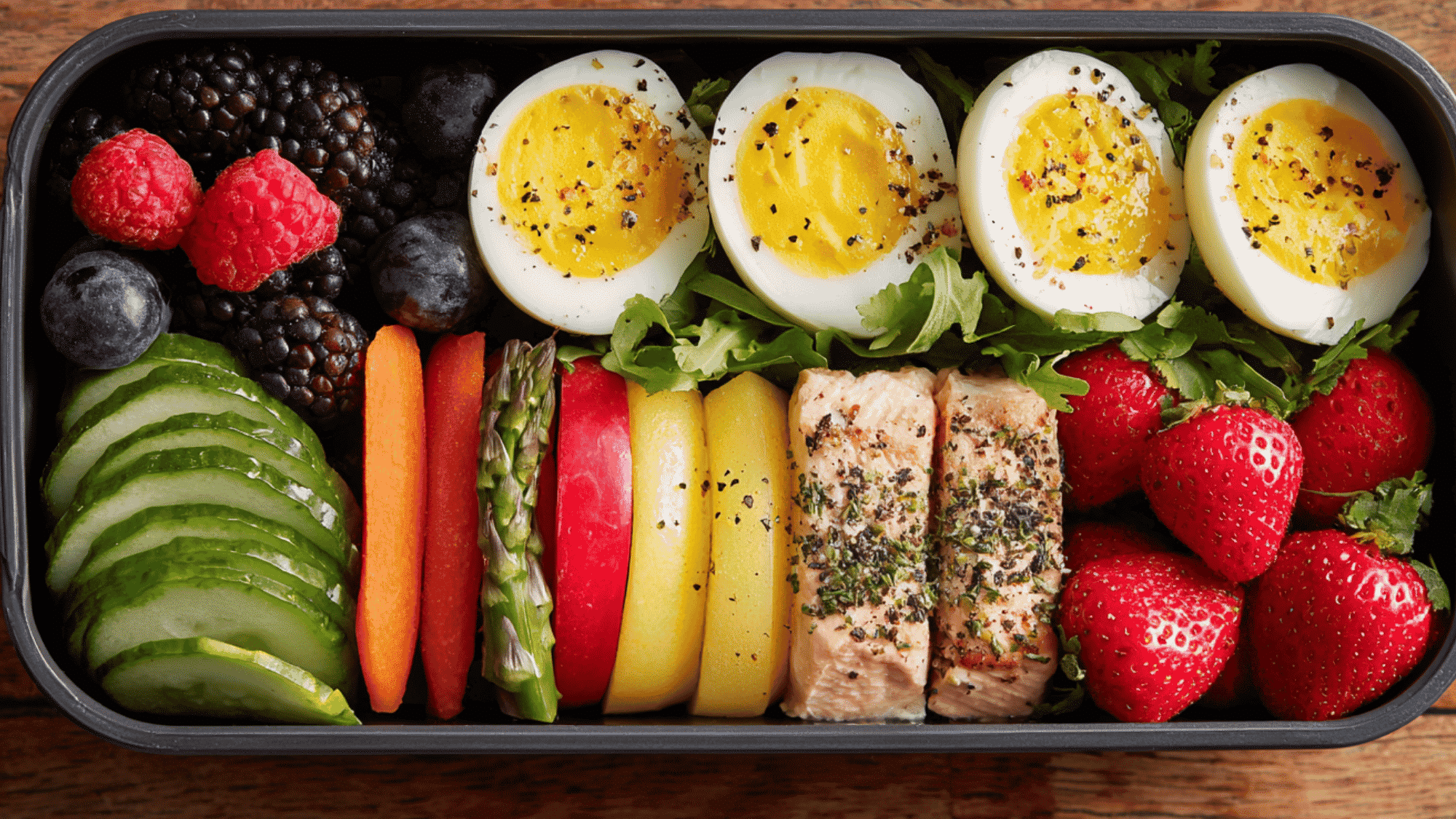 A bento-style meal prep lunch box with an egg, cheese, crackers, carrots, hummus, and fruit.