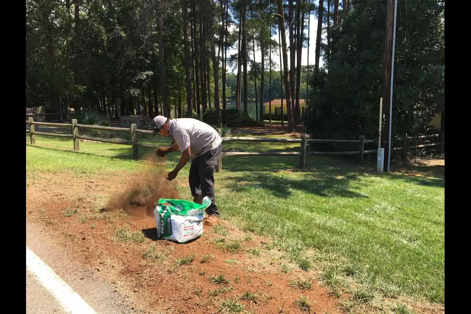 Person spreading grass seed and soil outdoors near wooden fence in sunny park