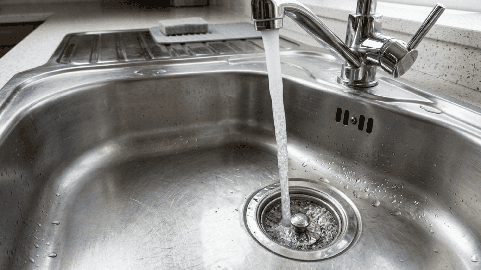 A hand rinsing a stainless steel sink with warm water from a faucet, clearing food debris and soap residue.