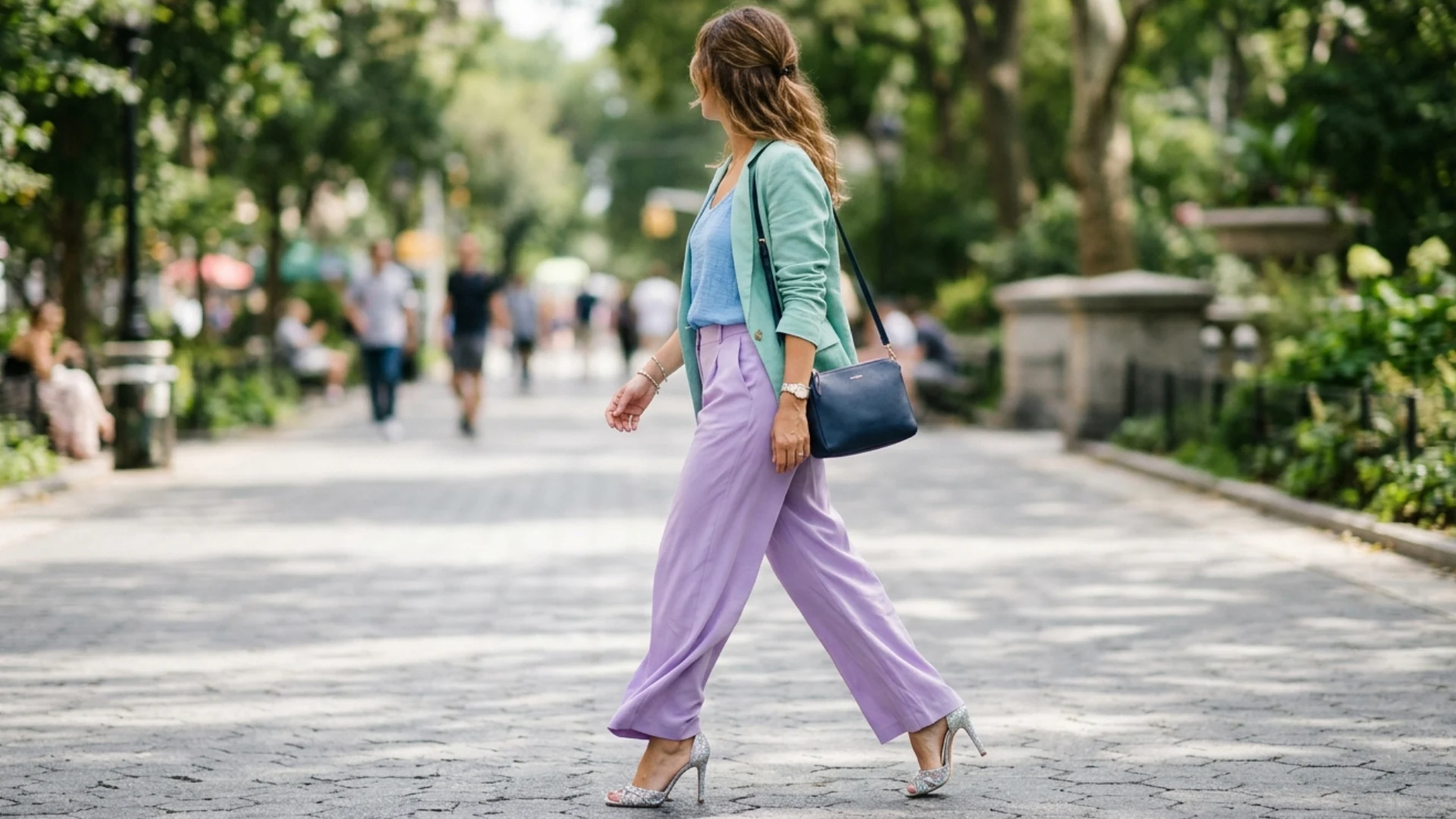 A woman in lavender pants and a teal jacket walks through a sunlit park. She carries a black bag, exuding confidence on a lively, tree-lined path.