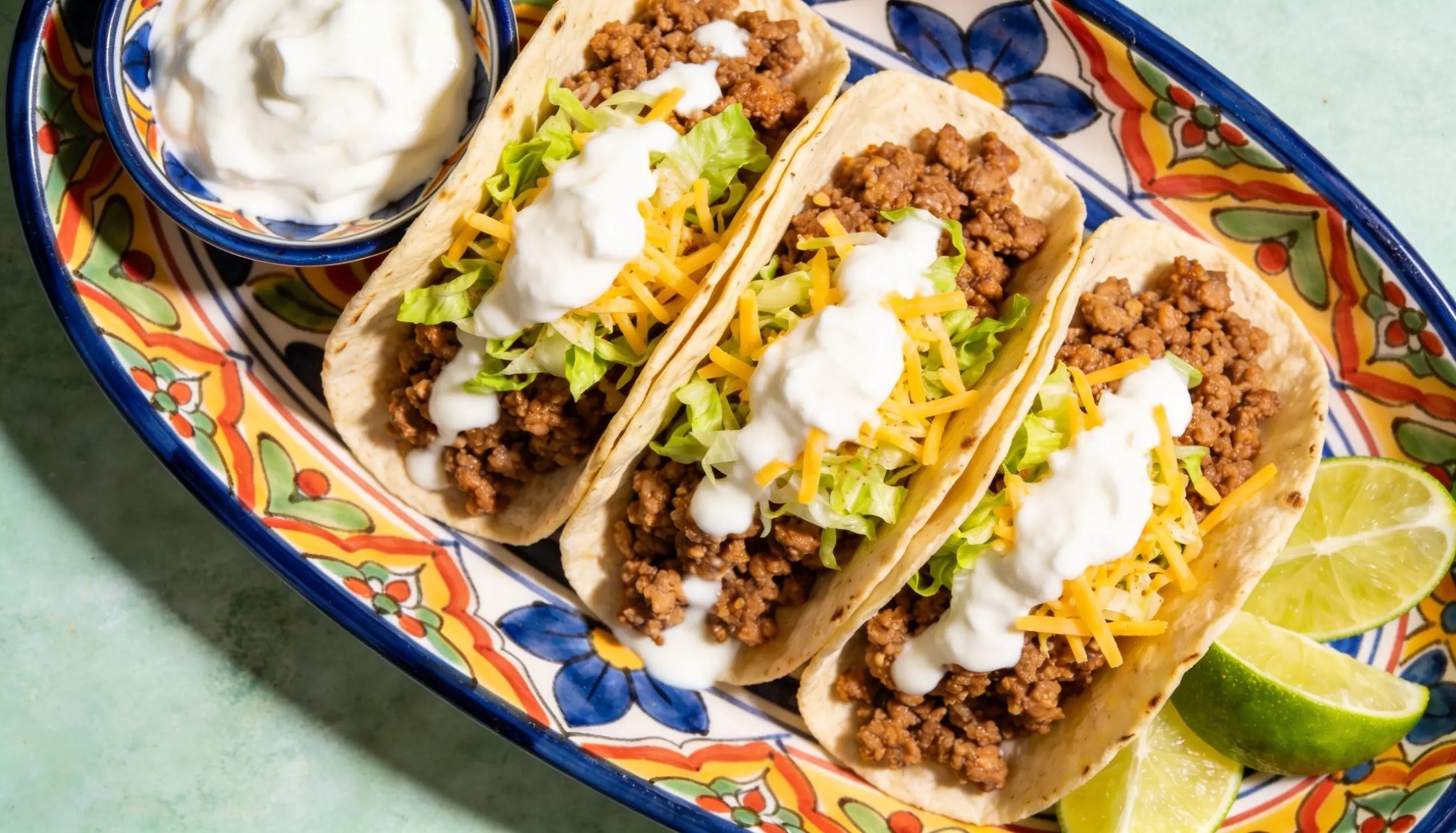 Three tacos filled with ground meat, shredded lettuce, cheddar cheese, and sour cream are arranged on a patterned plate with lime wedges and a small bowl of sour cream on the side.