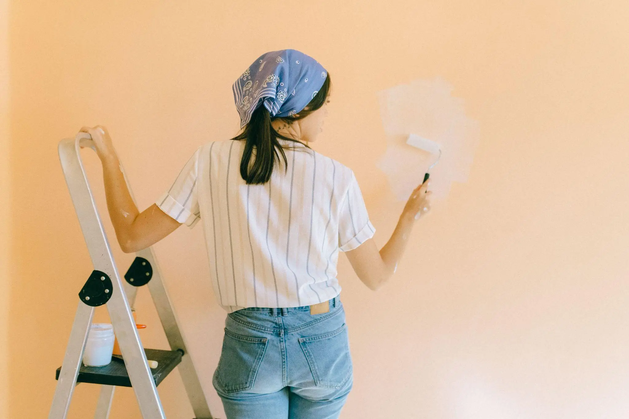 Person painting peach-colored wall with roller while standing on ladder in bright room