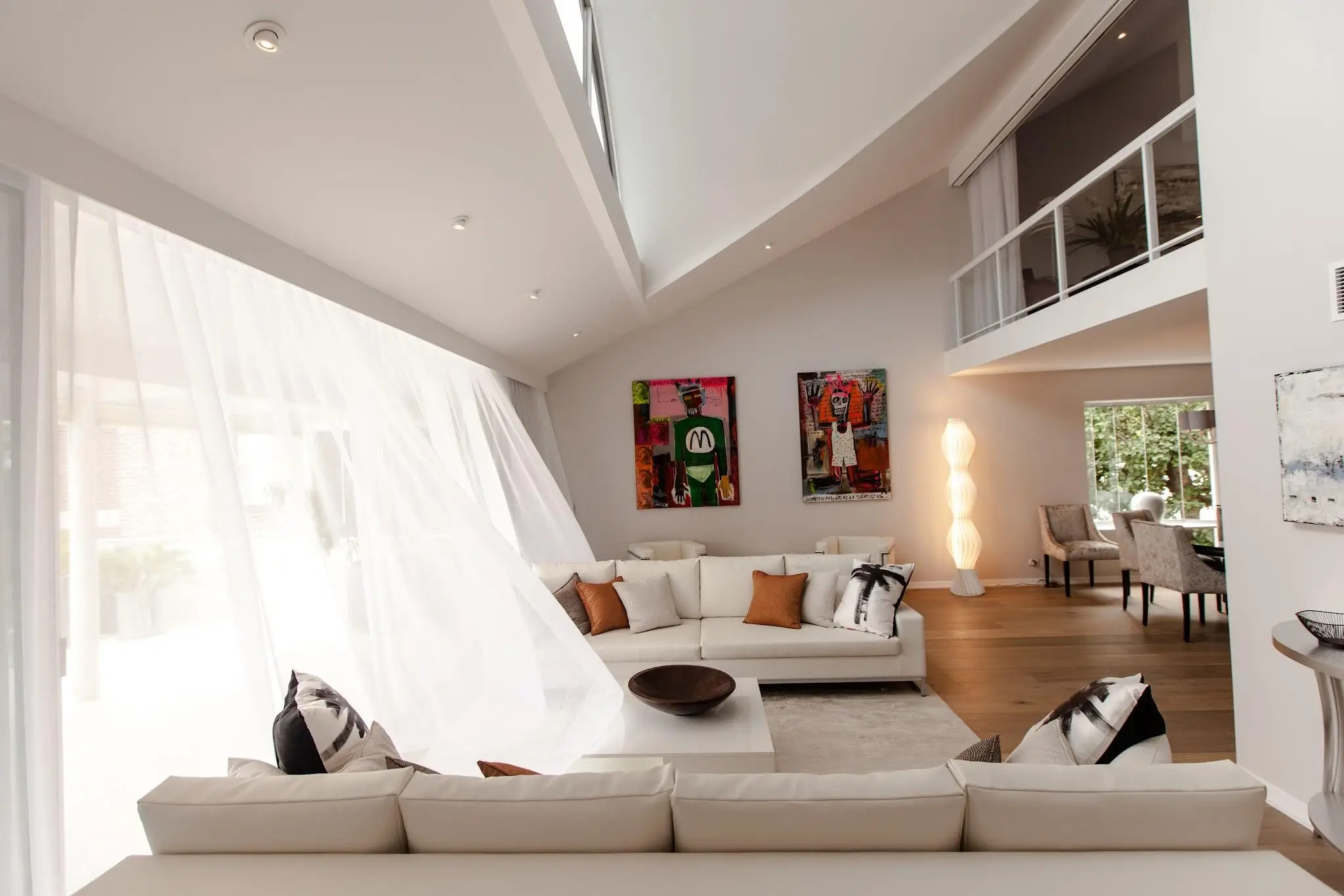 Spacious modern living room with white sofas, abstract art, and tall windows with sheer curtains