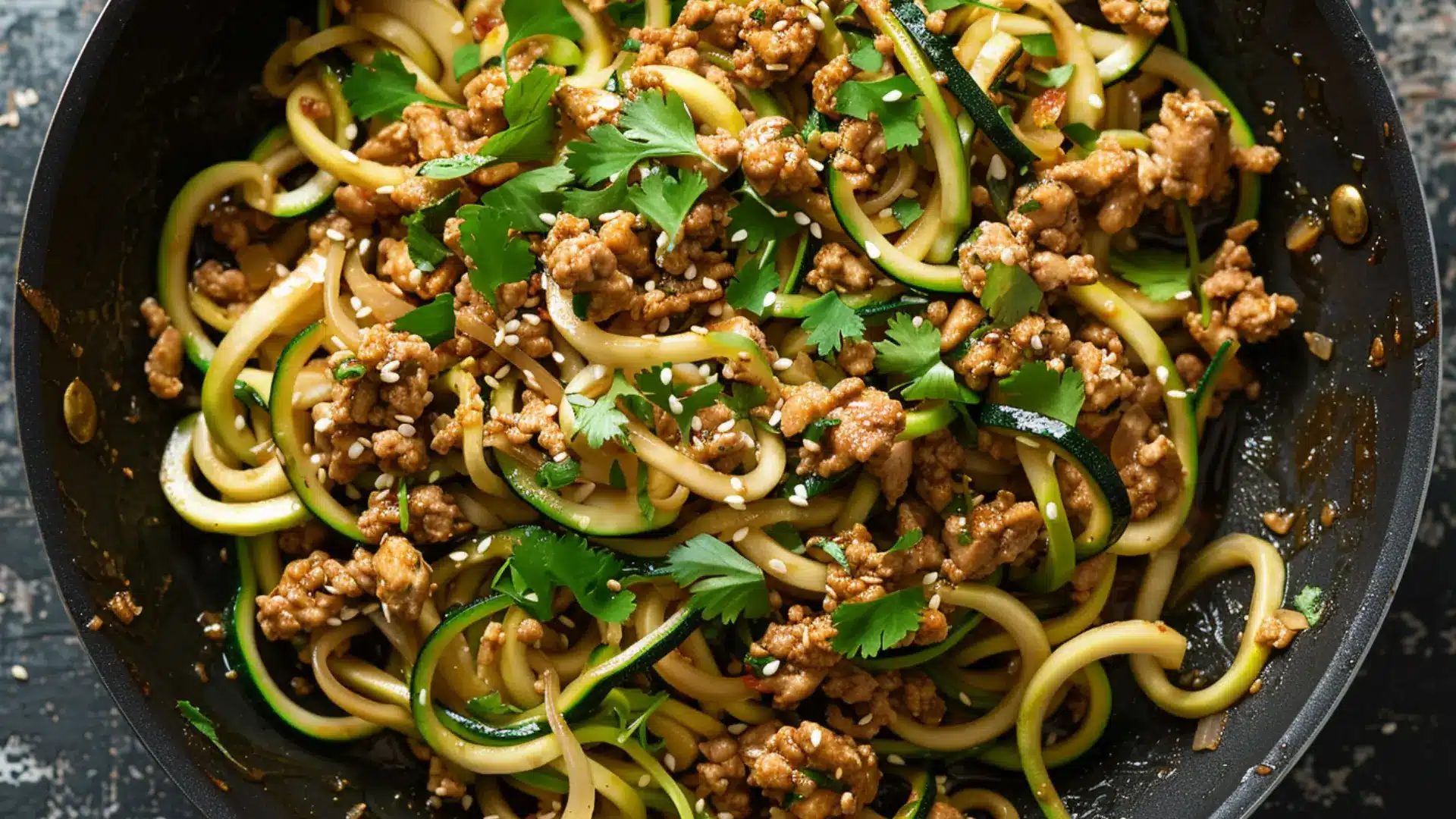Center focused ground chicken with garlic onion and spiralized zucchini noodles in skillet garnished with sesame seeds and chopped cilantro