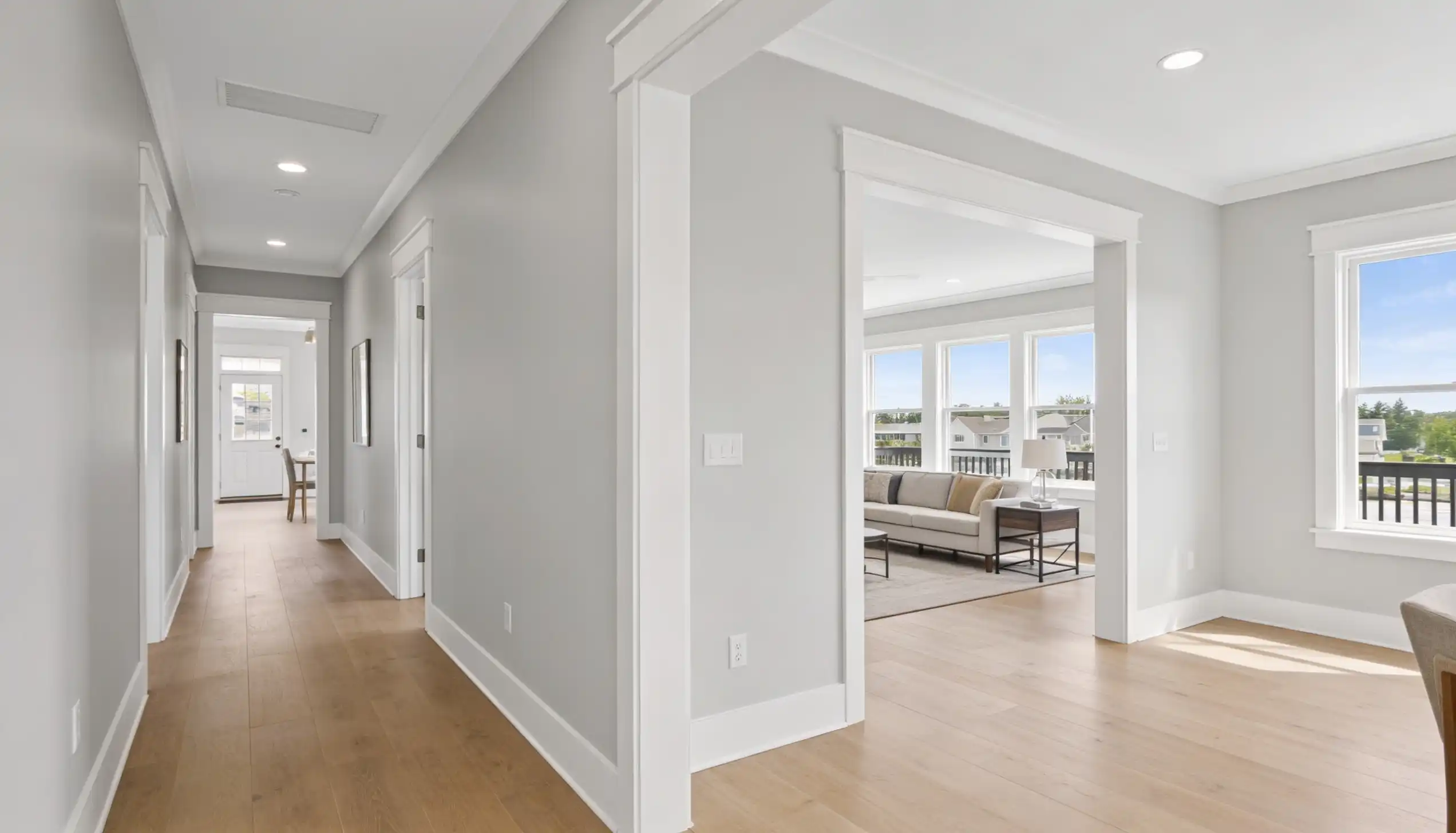 Bright, modern hallway with hardwood floors leading toward an open entryway into a living room featuring large windows and a light sofa.