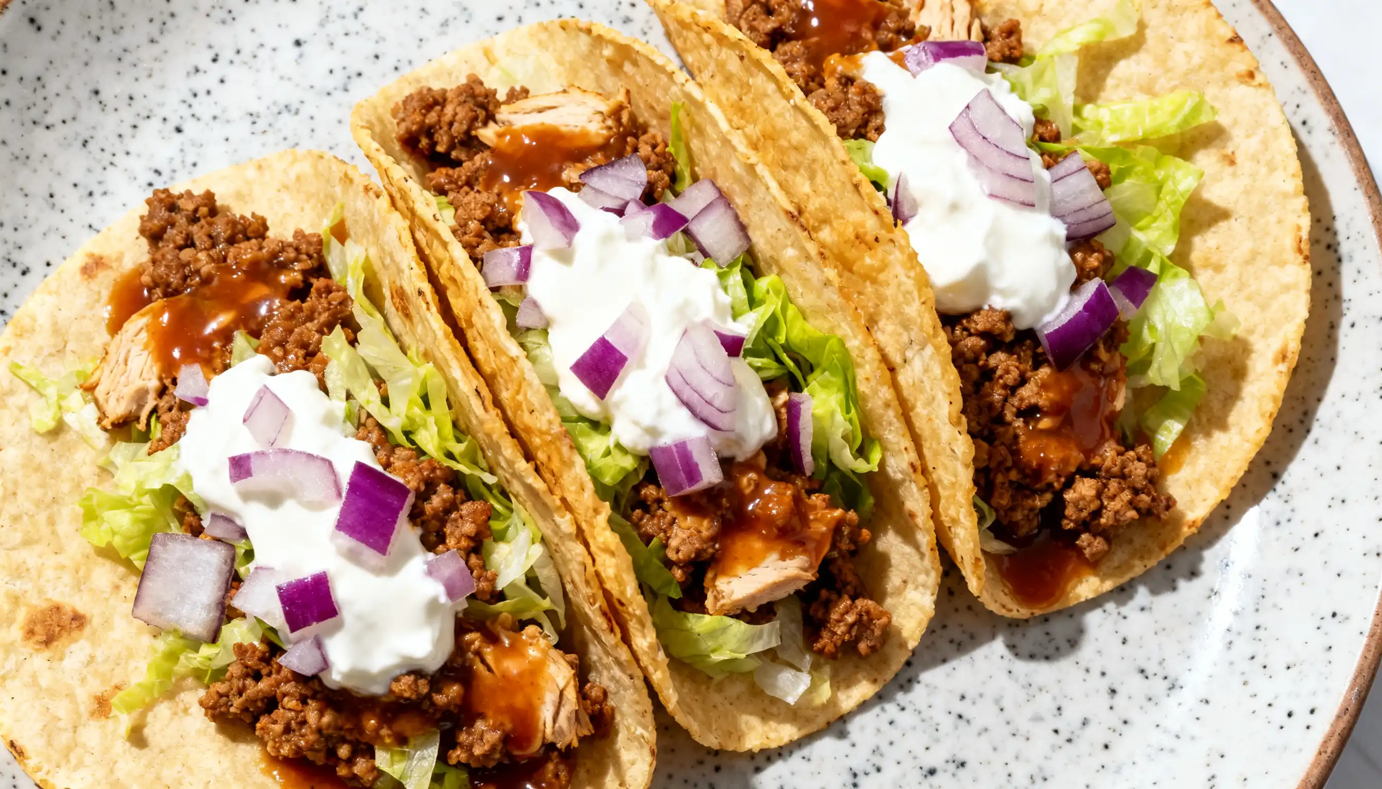 Three turkey tacos on a plate filled with ground meat, shredded lettuce, diced red onion, sour cream, and sauce in corn tortillas.