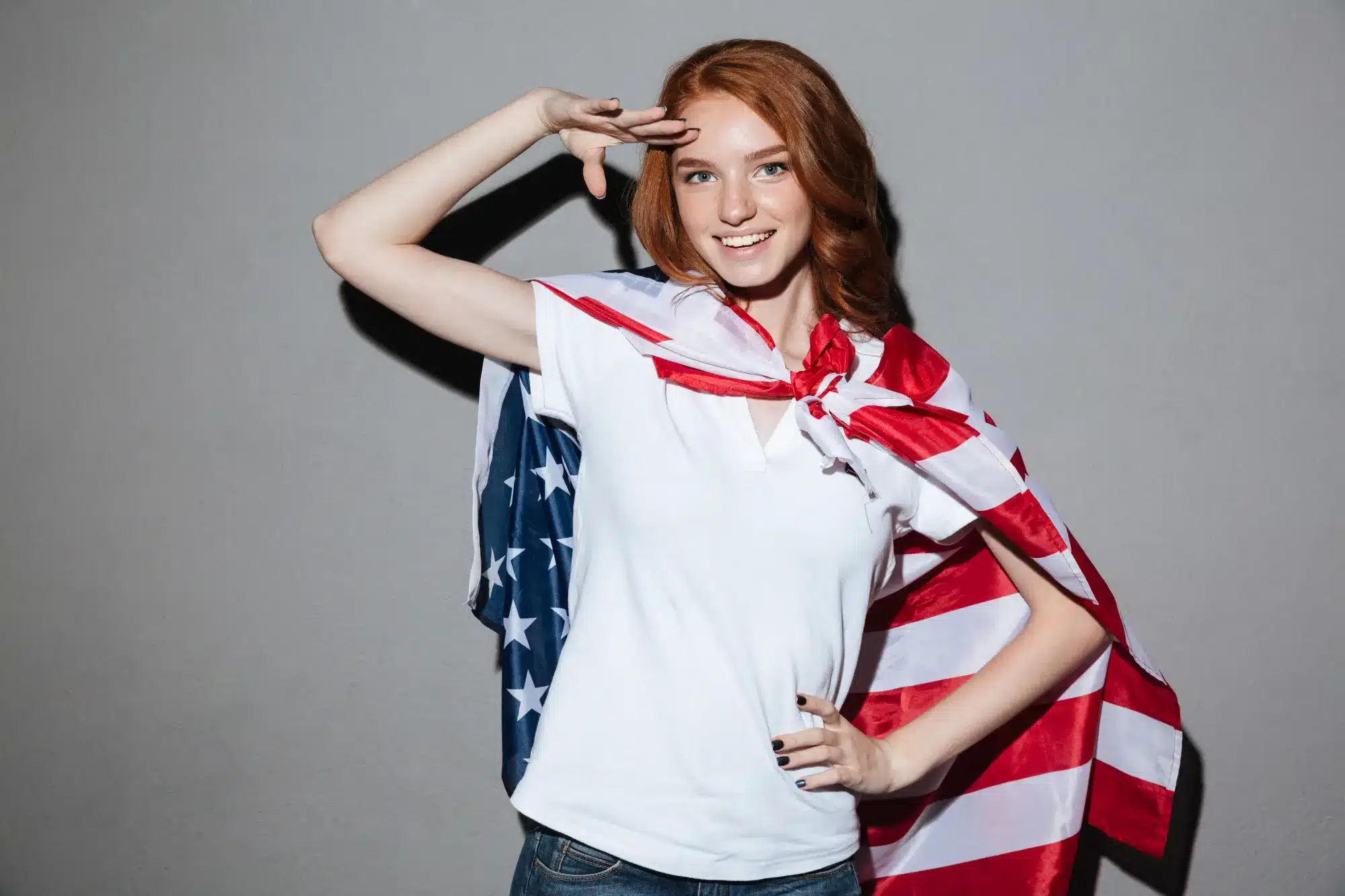 Young woman with American flag draped over shoulders saluting against gray wall
