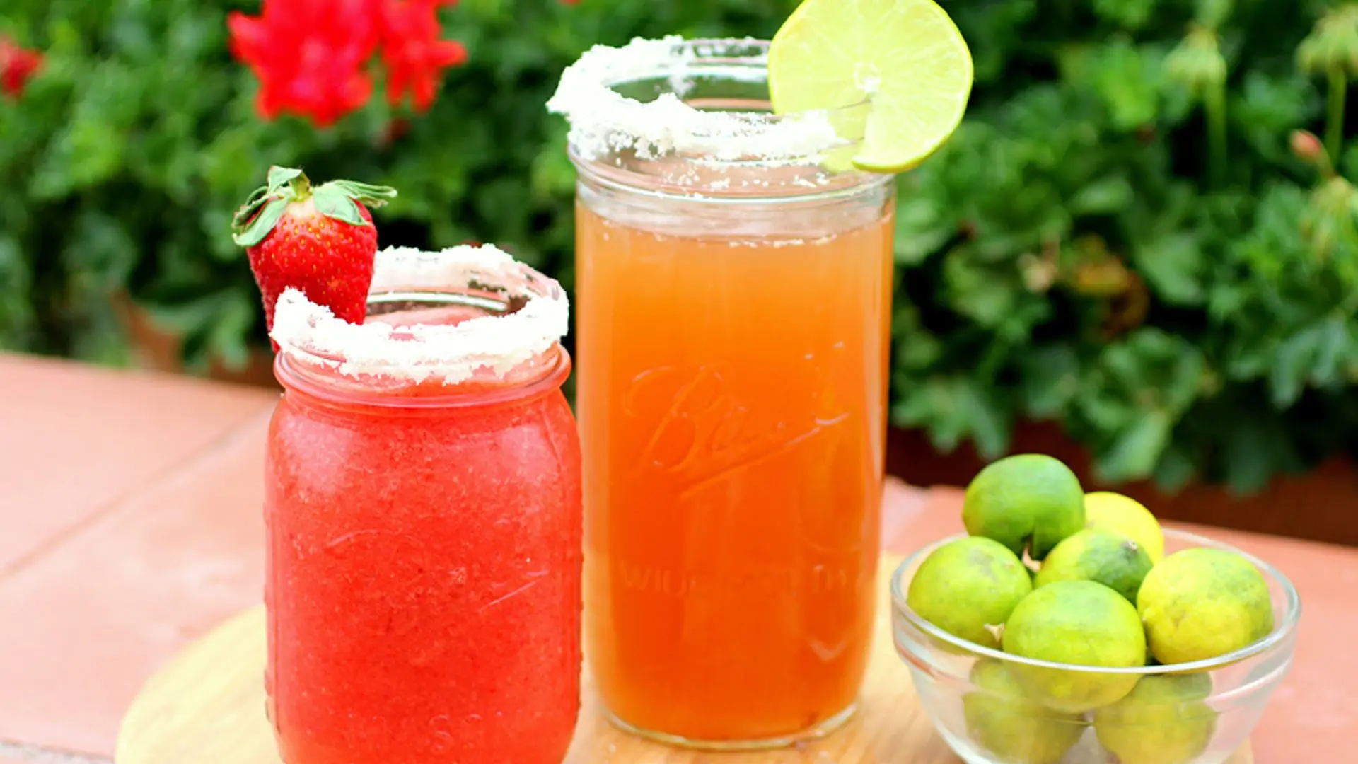 Two mason jar drinks, one strawberry and one orange, are garnished with salt rims, fruit, and lime slices next to a bowl of limes.