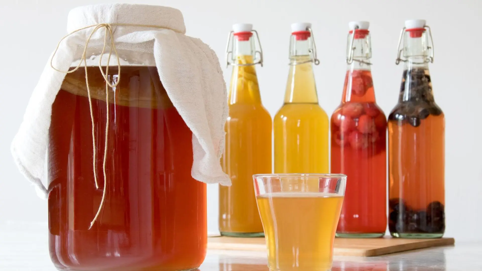 A large jar of kombucha with a SCOBY covered by cloth, next to bottles of flavored kombucha and a glass of the beverage.