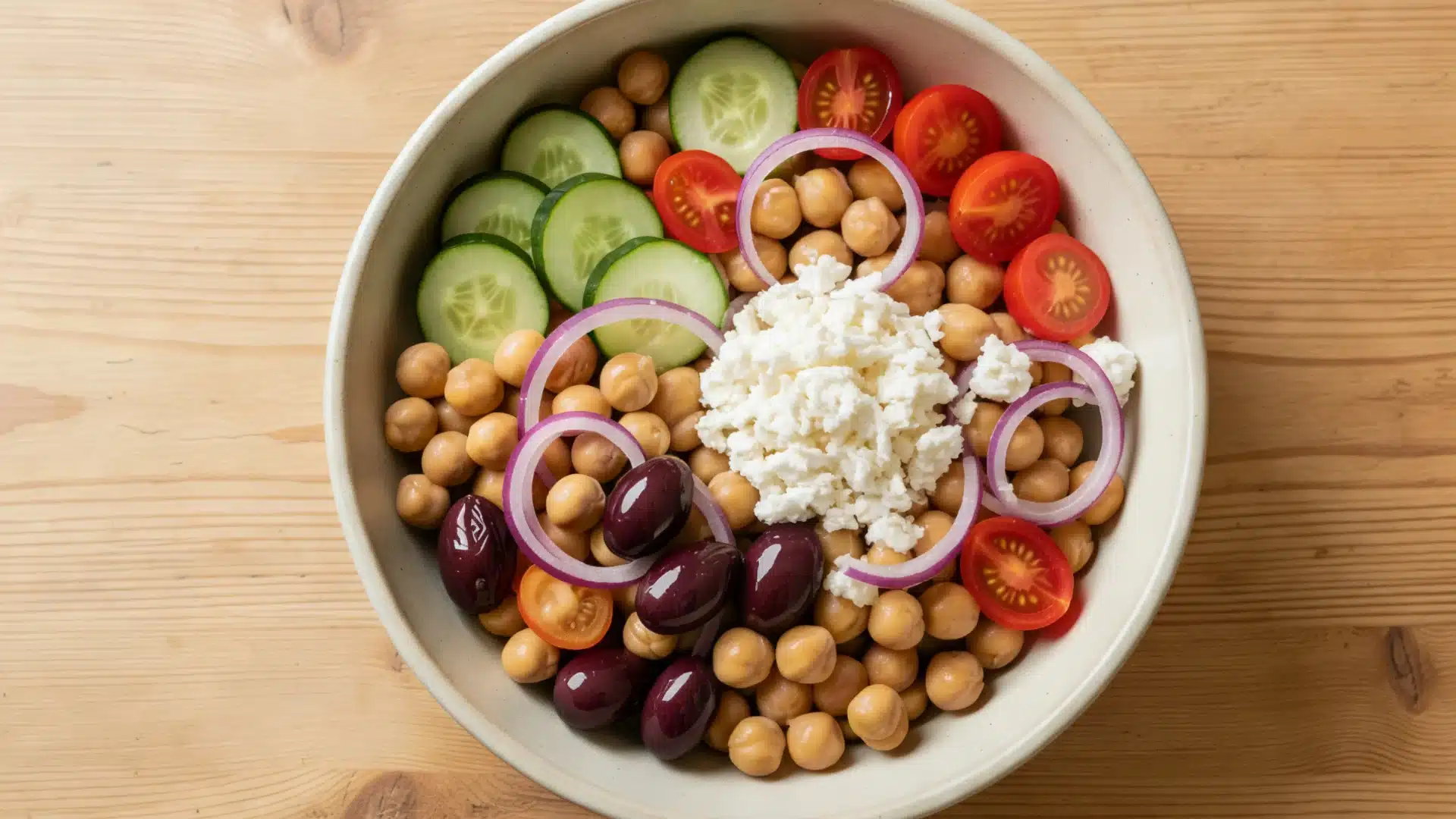 A bowl of chickpea salad with cucumbers, cherry tomatoes, Kalamata olives, red onion rings, and crumbled feta cheese