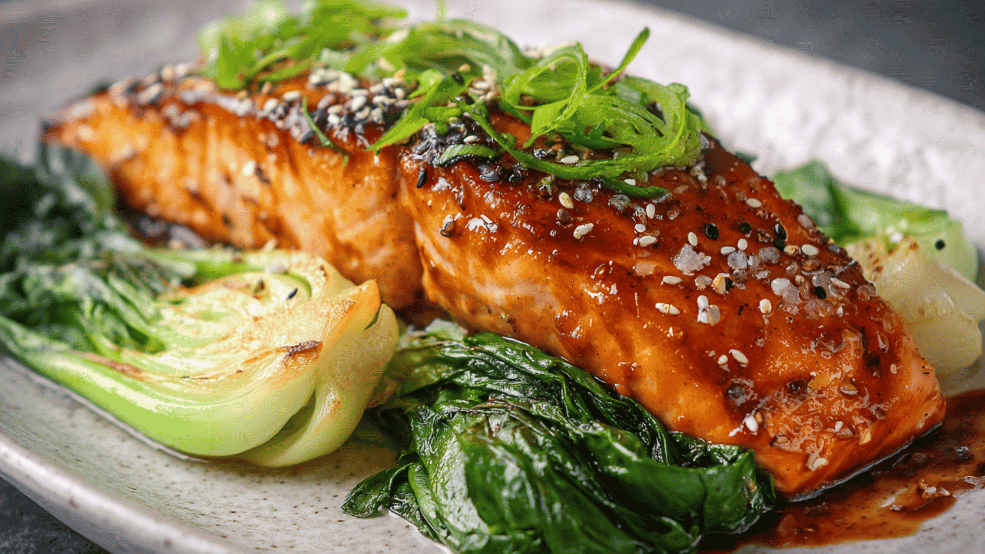 Two salmon fillets coated in a savory, caramelized white miso glaze, baked to perfection and served alongside crisp, vibrant green baby bok choy halves garnished with sesame seeds and sliced green onions.
