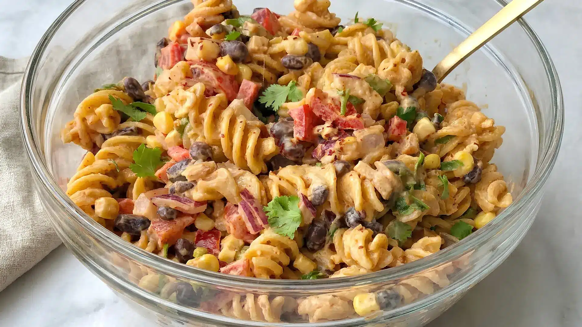 Colorful pasta salad in a glass bowl with rotini tomatoes cucumber bell pepper feta mozzarella and fresh herbs on a rustic kitchen counter