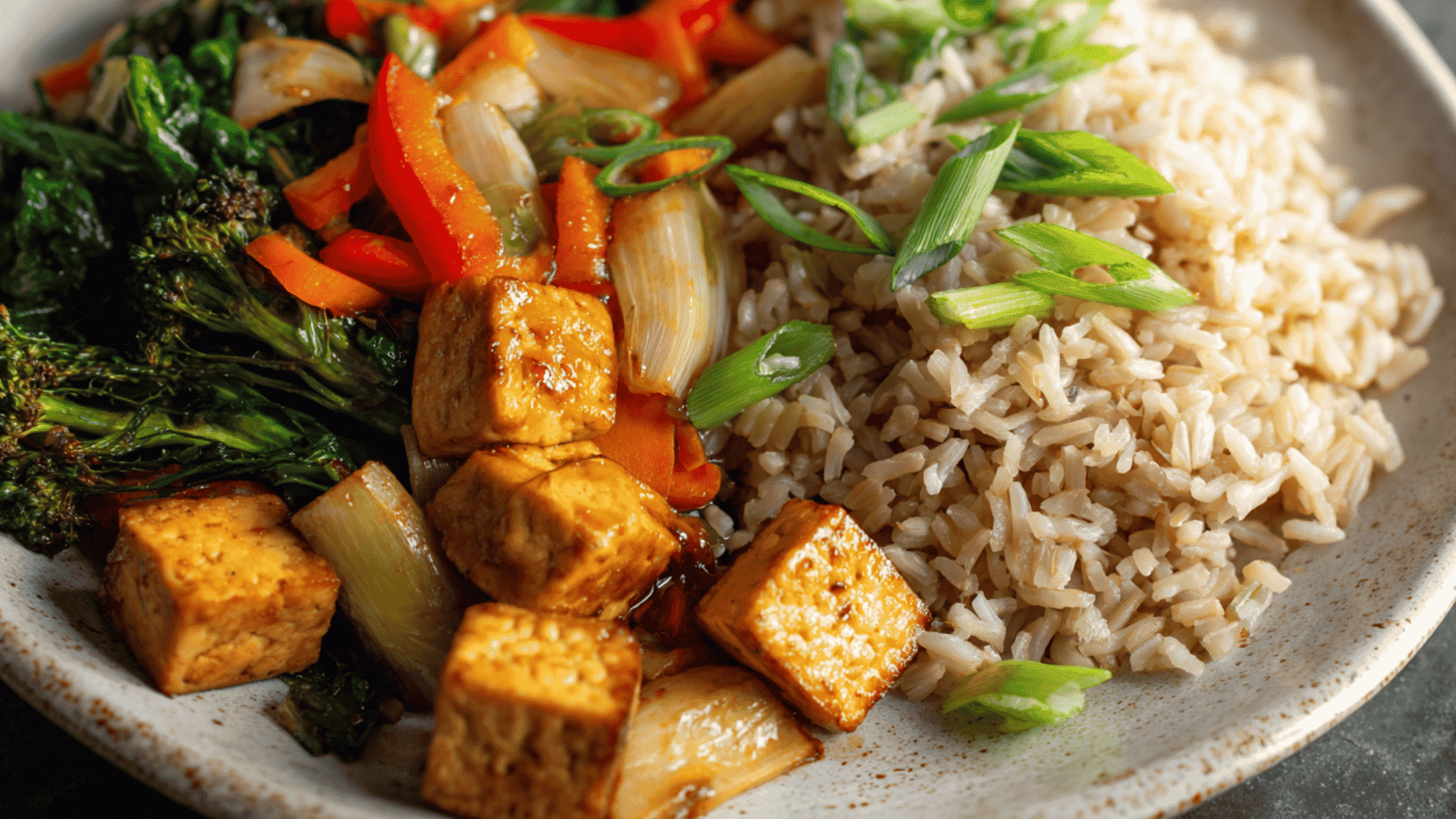 A vibrant plant-based tofu stir-fry served over fluffy brown rice in a meal prep container, featuring golden-brown cubed tofu, crisp green broccoli florets, and snap peas.