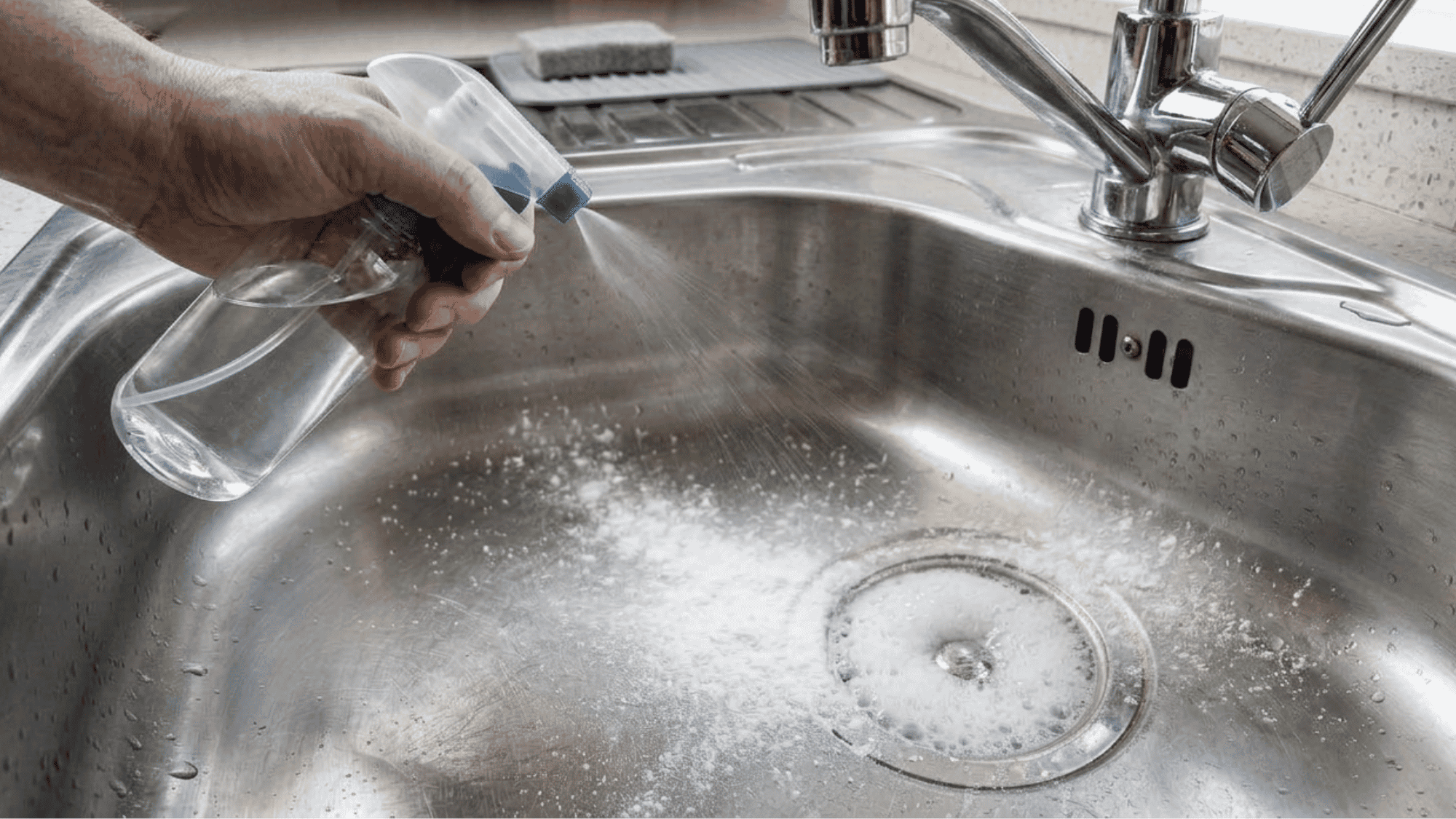 White vinegar being sprayed onto a stainless steel sink, creating a light fizzing reaction with the baking soda layer.
