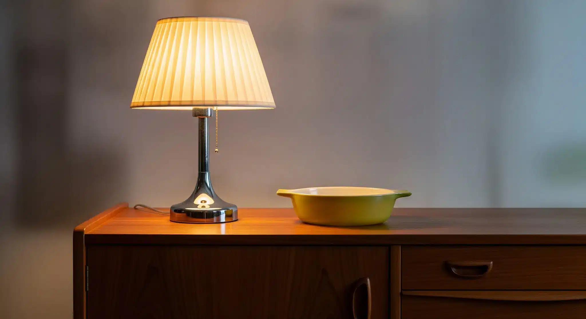 Lamp with pleated shade and yellow bowl on wooden sideboard in softly lit room
