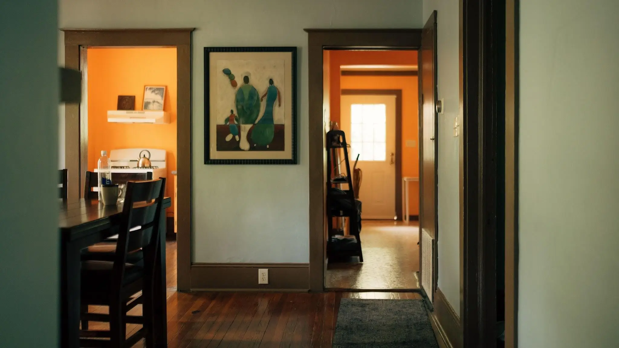 Dining table with chairs in foreground leading to an orange-walled kitchen and hallway with artwork