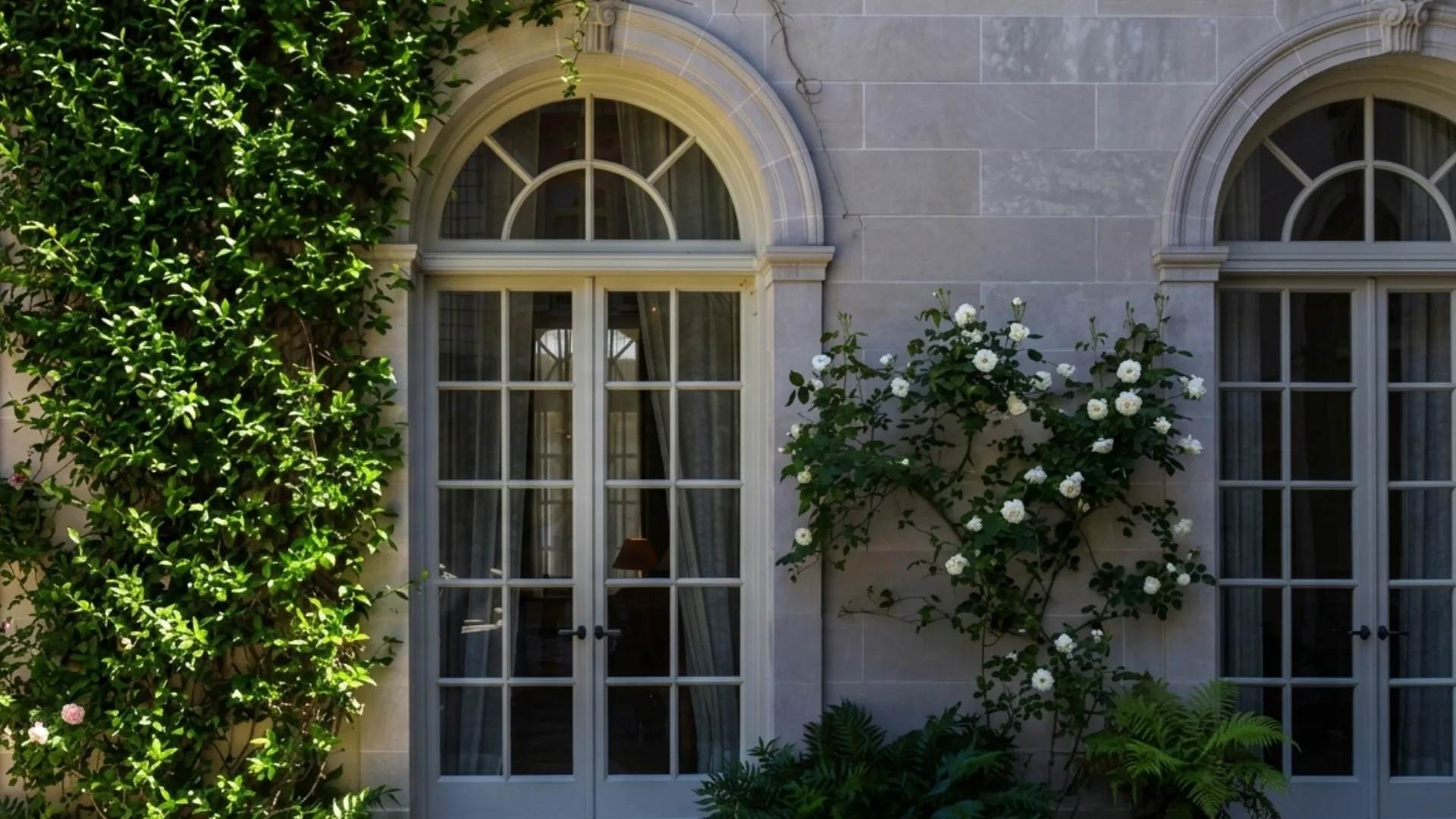 A pale stone wall features a pair of arched French doors and is overgrown with lush green vines and blooming white roses.