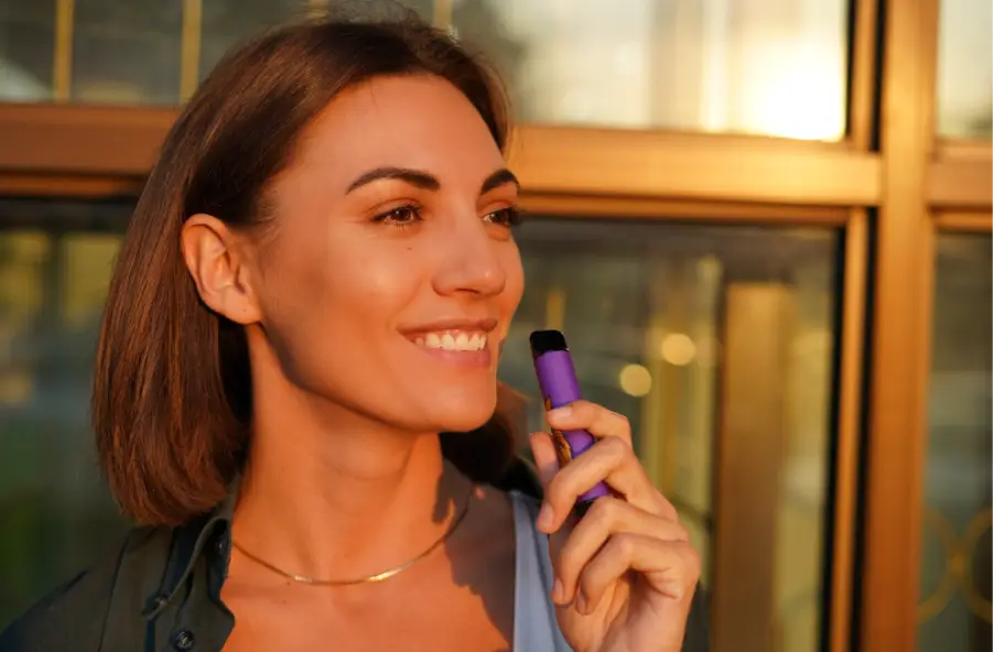 Smiling woman holding a purple vape pen in warm sunlight near glass window