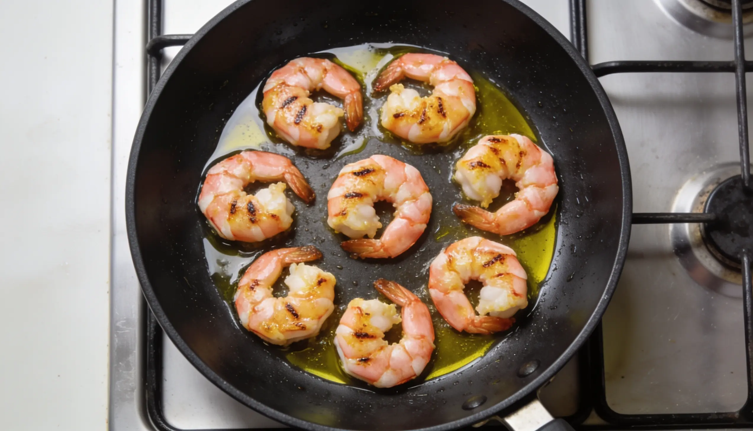 Seven large shrimp are pan-fried in olive oil on a stovetop.